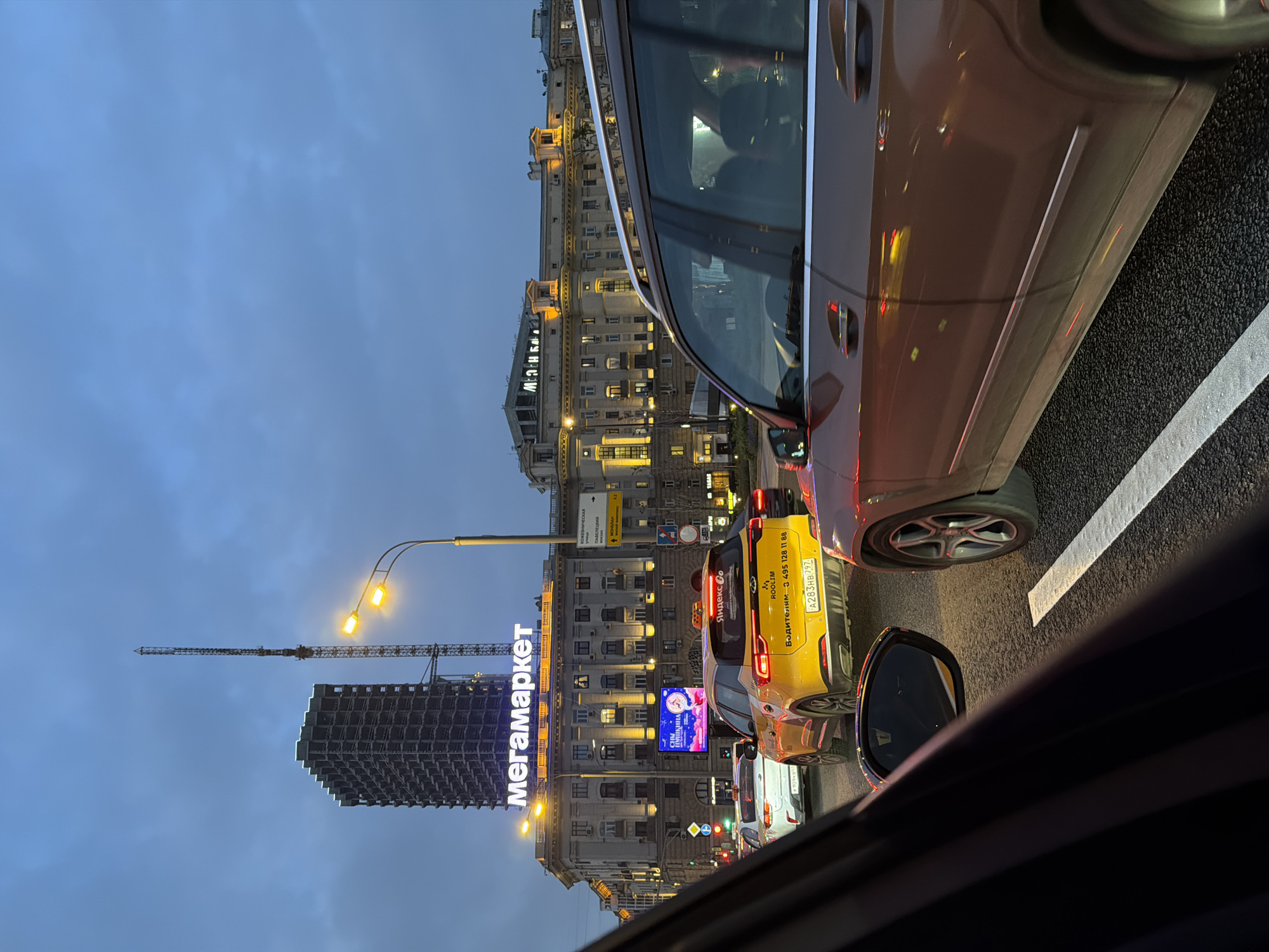 The image, taken from inside a car at dusk, shows a traffic jam in Moscow, Russia. In the foreground, the side of a brown car occupies a large portion of the right side, with a view of the road and a white lane marker visible. A yellow taxi is directly in front of the car, its license plate reading "A283HB 797" and displaying a phone number "3 495 128 11 88", and "Яндекс Со" on the back. Behind the taxi, other cars are visible in the traffic.
In the background, illuminated buildings line the street. A sign displaying "Мегамаркет" in bright neon letters is prominent on one of the buildings. To the left of the building, there is a construction site with a tall crane extending into the sky. A street lamp and building with the advertisement “Сны ПУШКИНА” and a sign with street names "КОЖЕВНИЧЕСКАЯ УЛИЦА" and "ПАВЕЛЕЦКАЯ ПЛОЩАДЬ" are also visible. The sky is a muted blue, indicating that it is dusk.