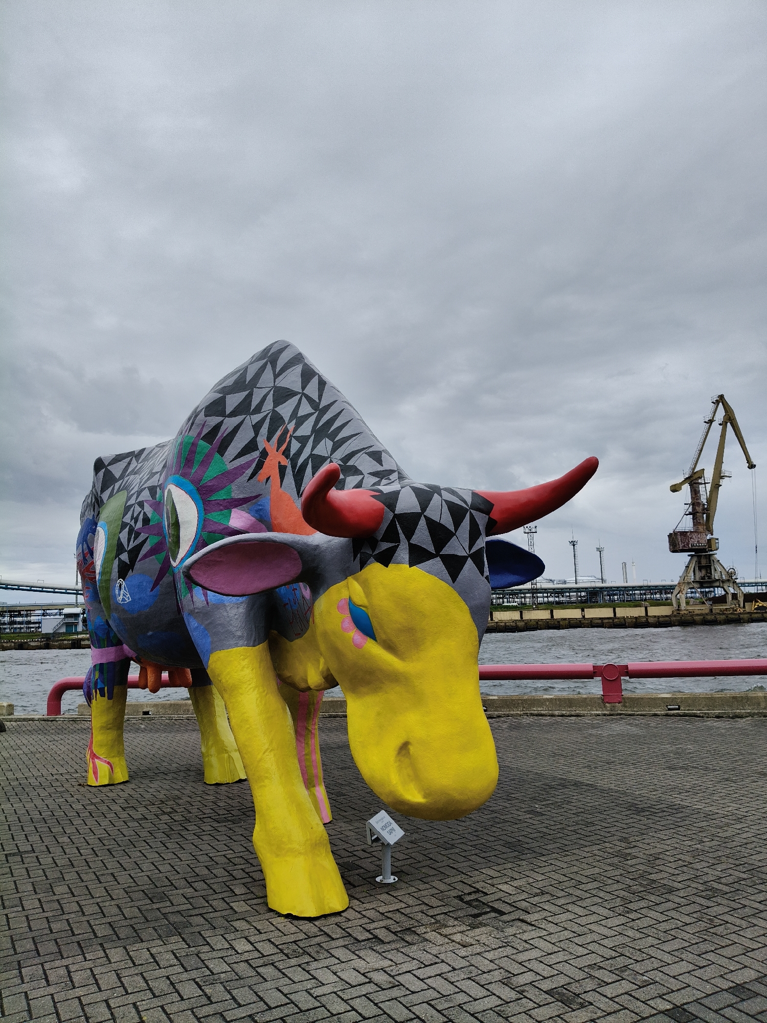 A large, brightly painted fiberglass sculpture of a cow stands on a paved walkway by a body of water. The cow is adorned with various geometric patterns and vibrant colors, including a black and gray triangular pattern on its back, yellow legs, red horns, and colorful designs on its body. In the background, industrial elements are visible, including a large yellow crane and a bridge structure, suggesting an industrial port or harbor area. The sky is overcast with gray clouds. This sculpture is located in the Ventspils city of Latvia.