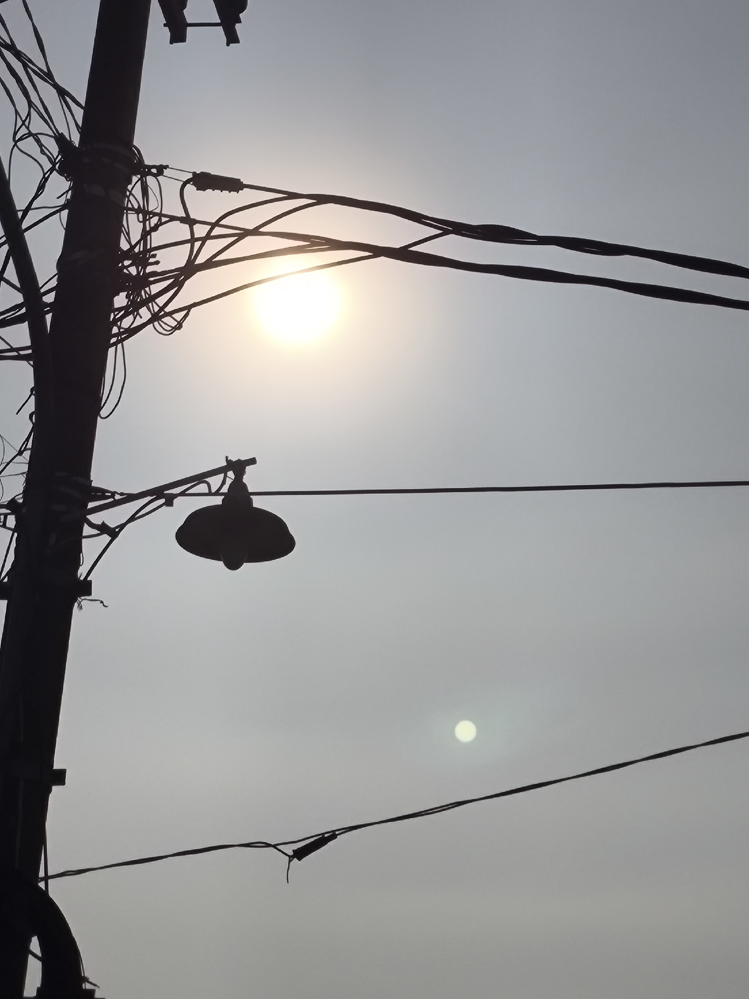 The image captures a dusky sky in Cilegon, Indonesia, with a prominent utility pole silhouetted against the fading light. A tangle of electrical wires emanates from the pole, crisscrossing the frame. Suspended from one of these wires is a streetlamp, its dome-shaped fixture casting a dark shadow. The sun, appearing as a bright, hazy orb, is visible near the upper left quadrant of the image, with a smaller, fainter disc, possibly another light source or a lens flare, appearing in the lower right. The overall atmosphere is dim and slightly overcast, suggesting either dawn or dusk.