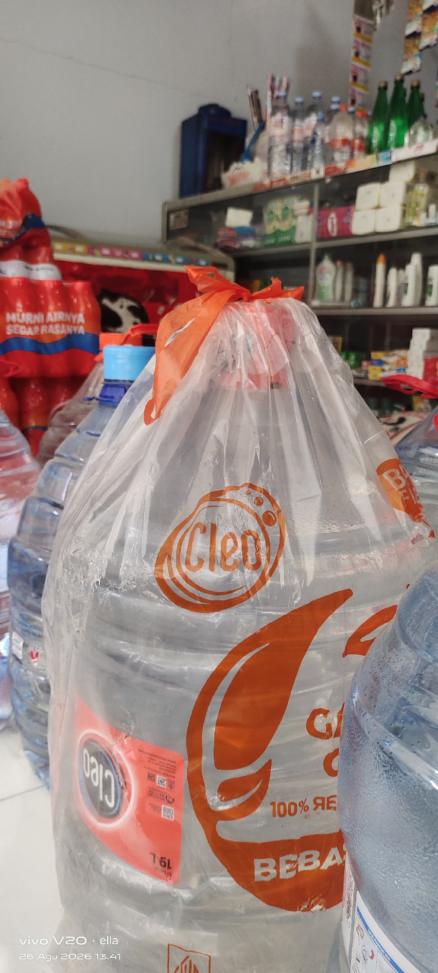 The image shows a close-up view of large plastic water containers inside what appears to be a small convenience store or warung. The primary focus is on a large, clear plastic container filled with water, covered with a transparent plastic bag that is tied with an orange ribbon at the top. The container has an orange logo with the word "Cleo" inside an oval shape, and orange leaf-like graphics. Below the "Cleo" logo, text reads "100% RE" and partially visible text "BEBA". Another smaller sticker on the container has "19L" and Indonesian text. To the left, several smaller plastic water bottles with blue caps are visible, and to the far left, red and white labeled water bottles are stacked. In the background, shelves are stocked with various goods, including bottles of water, cleaning supplies, and other packaged items. The overall setting suggests an indoor retail environment. The image was captured on August 26, 2026, at 13:41, and the text at the bottom left corner indicates the phone model "vivo V20" and the name "ella". While the image is tagged to Simo, Indonesia, this location is not explicitly visible within the scene itself.