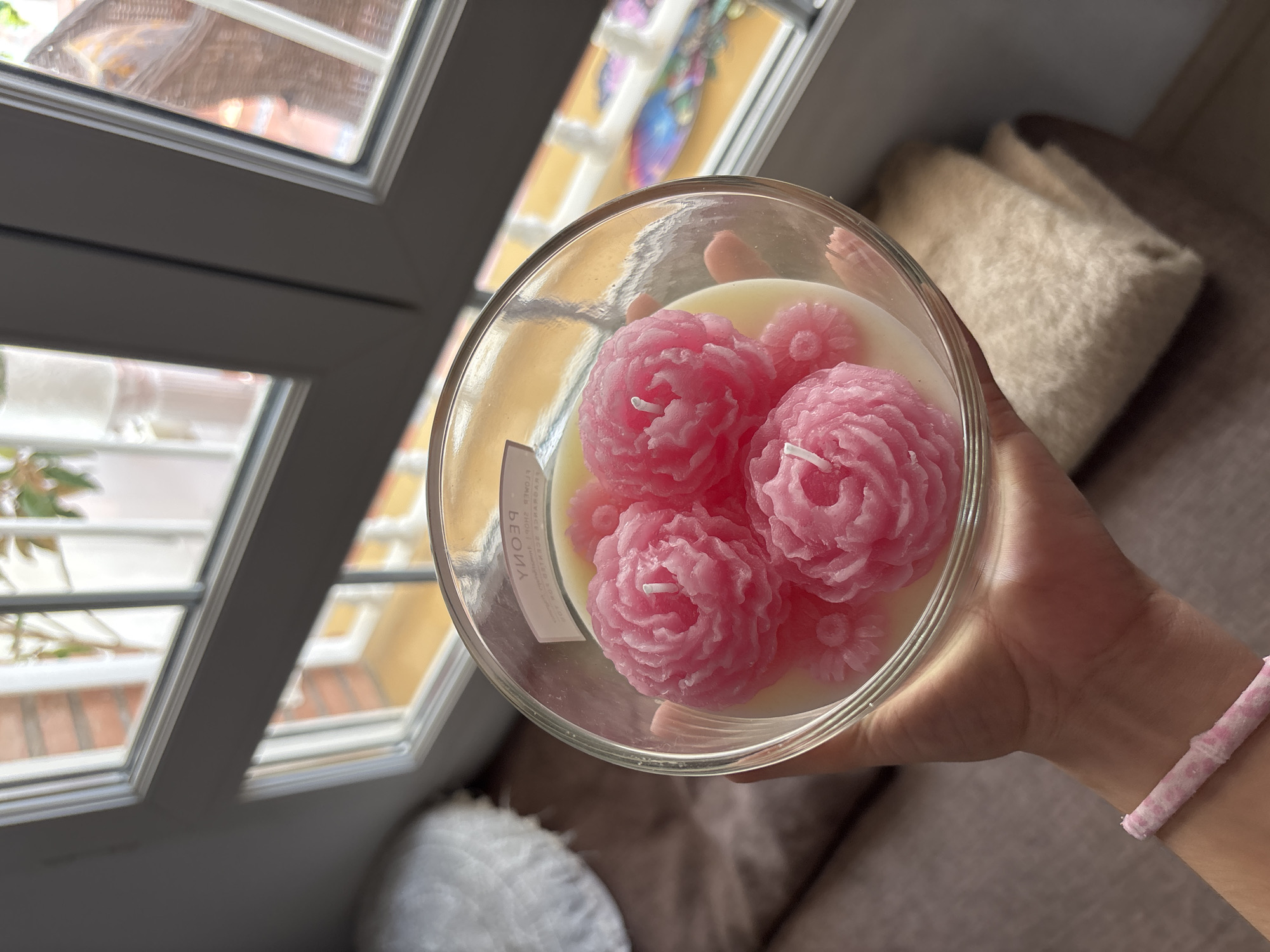 A hand holding a clear glass jar candle with pink peony-shaped wax flowers embedded in white wax. The candle is near a window with a view of brick buildings and greenery, suggesting an outdoor environment possibly in a residential area. The scene is lit by natural light.
