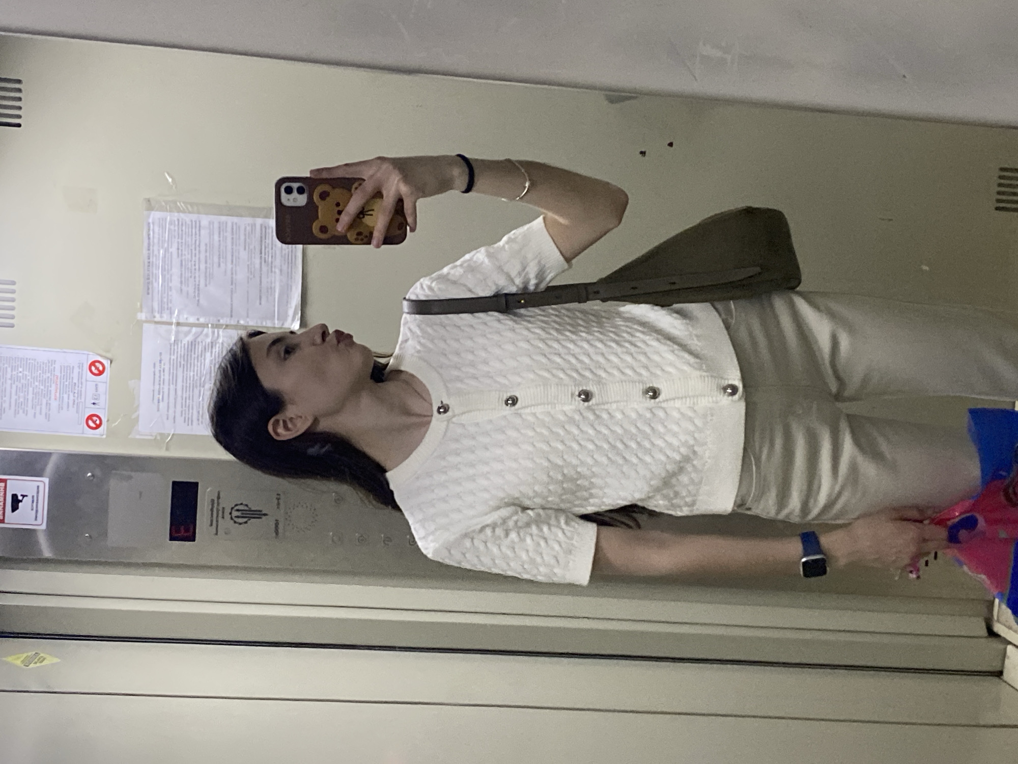 A woman is taking a selfie in a reflective elevator in Votkinsk, Russia. The elevator has a light beige interior, and the woman is standing to the right of the image. She is wearing a white textured sweater, light beige pants, and a blue smartwatch. She is holding a brown phone case with a bear design, and a green shoulder bag is draped across her body. The elevator features several notices and indicators, including safety signs, floor indicators (displayed as "3"), and other miscellaneous signs on the left side. The floor indicator shows the elevator is on the 3rd floor. The interior of the elevator appears well-lit and clean. Part of a blue plastic bag with pink contents is visible on the bottom right.