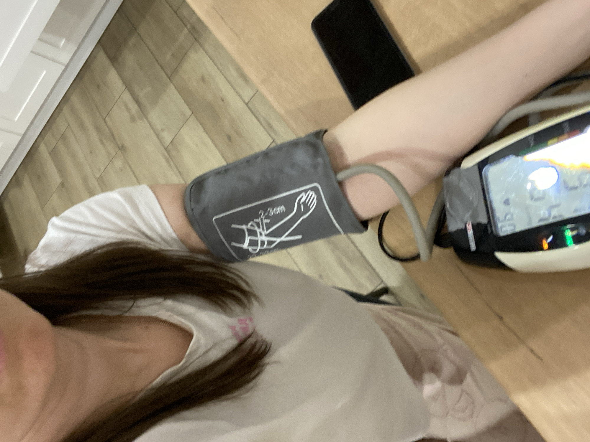 A person is taking their blood pressure in an indoor environment. A blood pressure cuff is wrapped around their left arm, and the digital blood pressure monitor is to the right of their arm. A black smartphone rests near the arm. The floor is made of wood-like planks. A white cabinet is visible in the upper-left corner of the image. The person is wearing a white t-shirt. This photo was taken indoors, but location in the city of Chaikovsky, Russia is unknown.