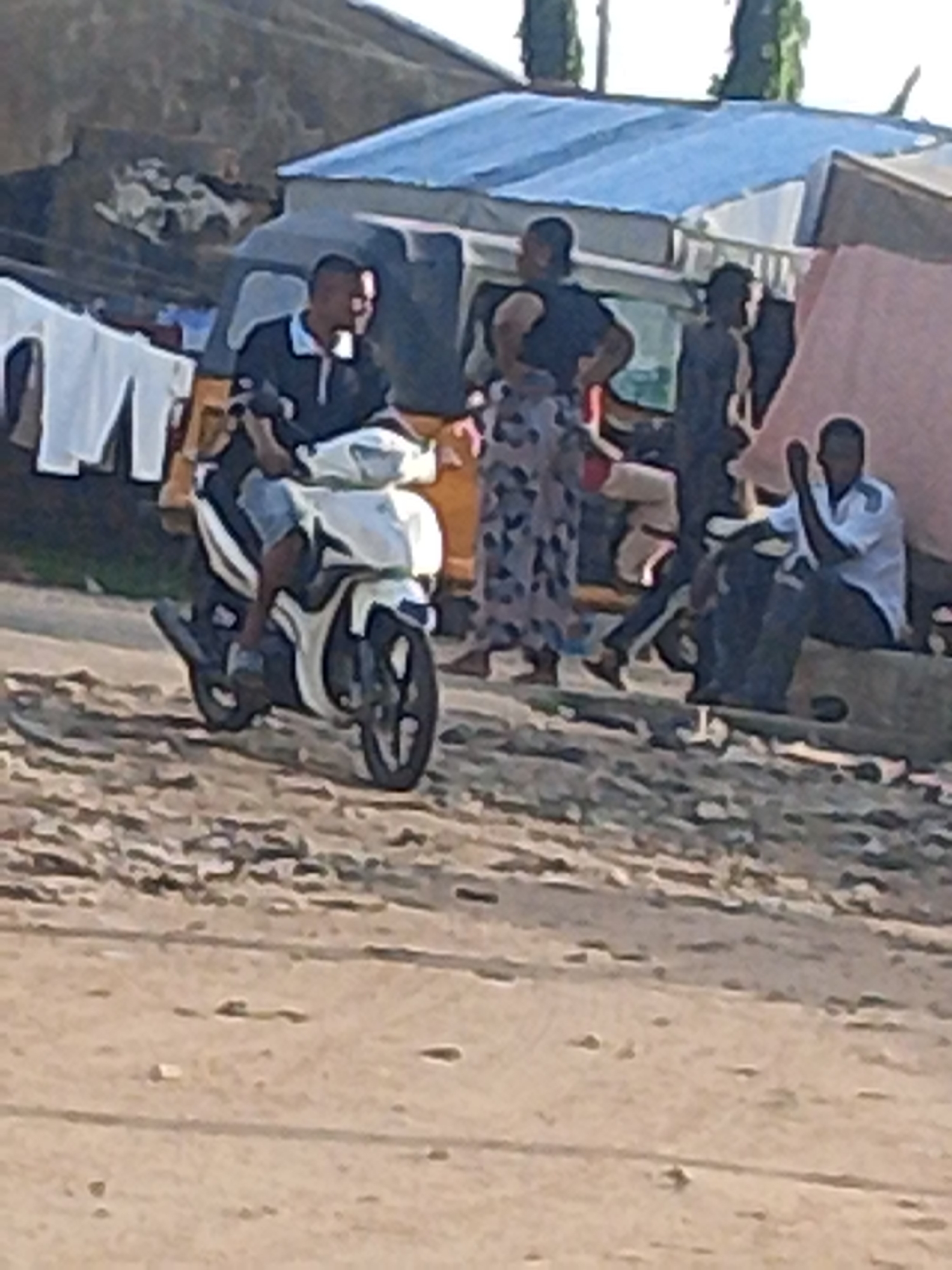 A man is riding a white and black motorcycle on a dirt road in Jalingo, Nigeria. He is wearing a dark polo shirt and blue shorts. Behind him, a white van is parked, and several people are standing around. One person, wearing a black vest and patterned pants, is looking towards the left. Another person, wearing a white t-shirt and dark pants, is sitting on a concrete block. A pink tarp is visible behind them. Clothes are hanging on a line to the left of the van. The environment appears to be a somewhat informal or commercial area, with rough terrain and simple structures.