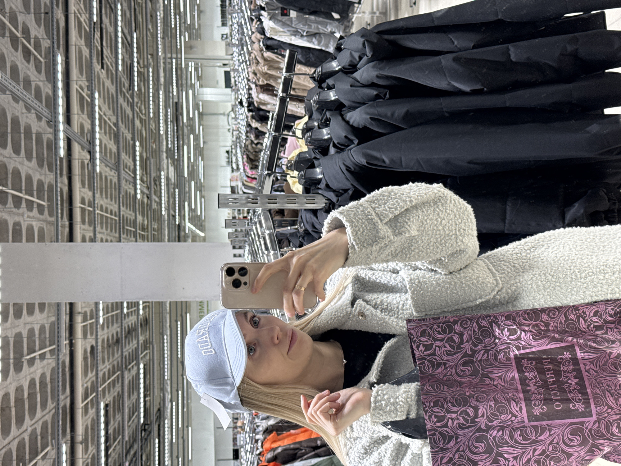 The photograph shows a woman with blonde hair and blue eyes, wearing a light blue baseball cap and a white, textured jacket. She is holding a beige smartphone in her right hand, presumably taking a selfie. She is also holding a dark purple shopping bag with an intricate pattern and the Cyrillic word "БЕВЕССЛО" printed on it.
The background reveals that she is in a large retail store. Clothes are hanging on racks behind her. The store features high ceilings with a grid of concrete walls and fluorescent lighting fixtures. The retail store is located in Izhevsk, Russia.