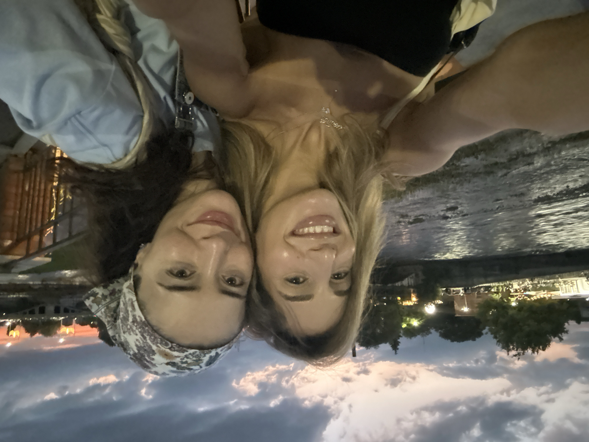 The image features a selfie taken by two women at dusk in Vladikavkaz, Russia. The women are positioned in the foreground, their faces close together and smiling. The woman on the left has dark hair and is wearing a light blue denim jacket and a patterned headscarf. The woman on the right has blonde hair and is wearing a black top and a delicate necklace.
The background depicts a waterfront scene. On the left, a structure with railings, possibly a bridge or pier, is visible. To the right of the women, a body of water reflects the lights of a distant cityscape. The city lights appear as a warm glow against the darkening sky. Trees are interspersed throughout the cityscape.
The sky is filled with dramatic clouds, indicative of twilight. The overall lighting suggests the sun has recently set, casting a soft, diffused light over the scene. The mood appears joyful and relaxed, as evidenced by the smiles of the subjects. The image is taken from a low angle, with the women's heads pointing upwards. There is no visible text in the image.