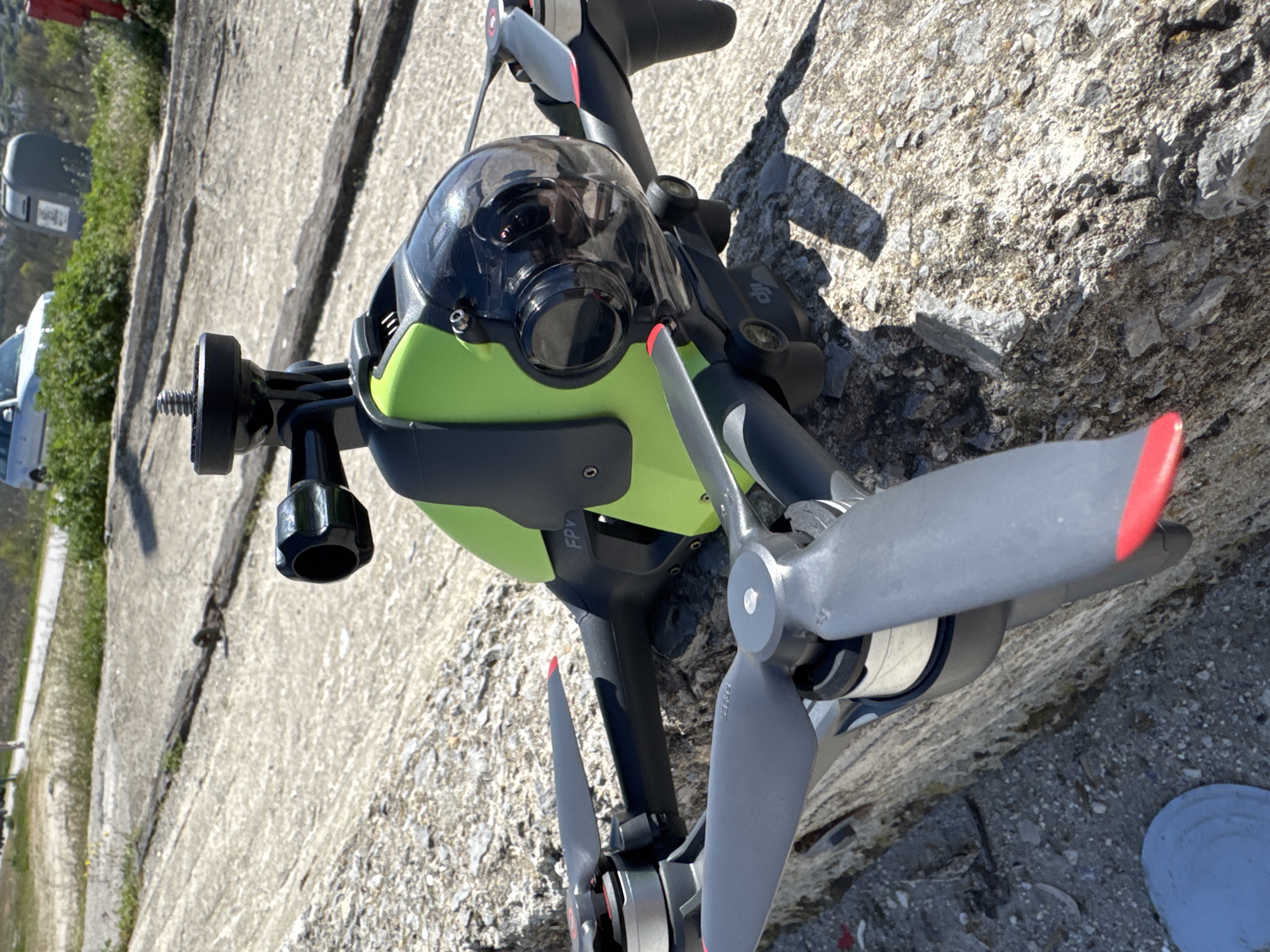 The image displays a DJI FPV drone resting on the ground under bright daylight. The drone features a striking neon green body with a black and grey frame, and large grey three-bladed propellers tipped with red. A transparent protective dome covers the main camera, and reflections of the surrounding environment, including what appears to be a person's hands holding a phone (likely the photographer), are visible on its surface. A black mounting bracket with a threaded bolt is attached to the drone's front left side, suggesting an accessory mount.
Visible text on the drone includes "FPV" on one of its arms and a small "DJI" logo near the camera. The setting is an outdoor location with strong sunlight, casting distinct shadows. The ground surface is varied, comprising textured concrete or paved sections alongside rougher, gravelly areas. In the background, there is green vegetation, possibly bushes or trees, and hints of distant objects, including a white vehicle and a dark, box-like structure. A grey, circular object is partially visible in the lower right foreground. The scene suggests an open, natural, or semi-urban outdoor area, consistent with a coastal city like Varna, Bulgaria. The drone appears to be stationary, possibly having landed or awaiting use. The entire image is oriented vertically (rotated 90 degrees counter-clockwise).