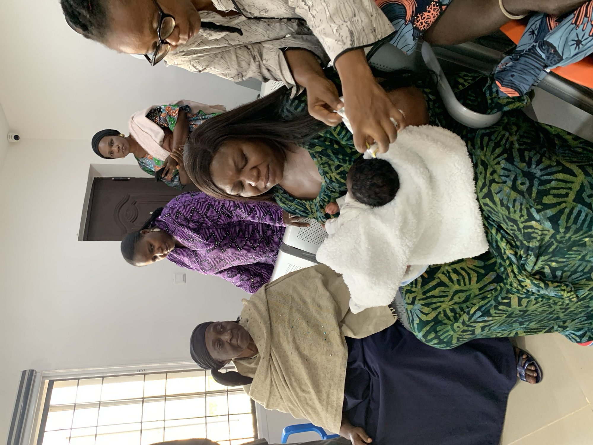 An image taken indoors at a location in Jos, Nigeria, shows a group of women and a newborn baby. The environment appears to be a waiting area, with white walls, a security camera on the ceiling, and a window with blinds.
In the foreground, a woman in a green and black patterned dress is holding a newborn baby wrapped in a white towel. Her eyes are closed, and she appears to be in a moment of quiet contemplation. To her right, another woman with glasses is looking down, her hands occupied with what appears to be assisting with the baby.
Behind them, an elderly woman is seated, wearing a light brown shawl over a dark blue dress. She is looking towards the front with a thoughtful expression. Two other women are visible in the background. One is wearing a vibrant purple patterned dress, and the other is in a colorful outfit with a light pink scarf, holding a mobile phone.
The overall impression is of a private, familial moment, likely in a medical facility or a similar public space adapted for waiting. The lighting suggests it is daytime.