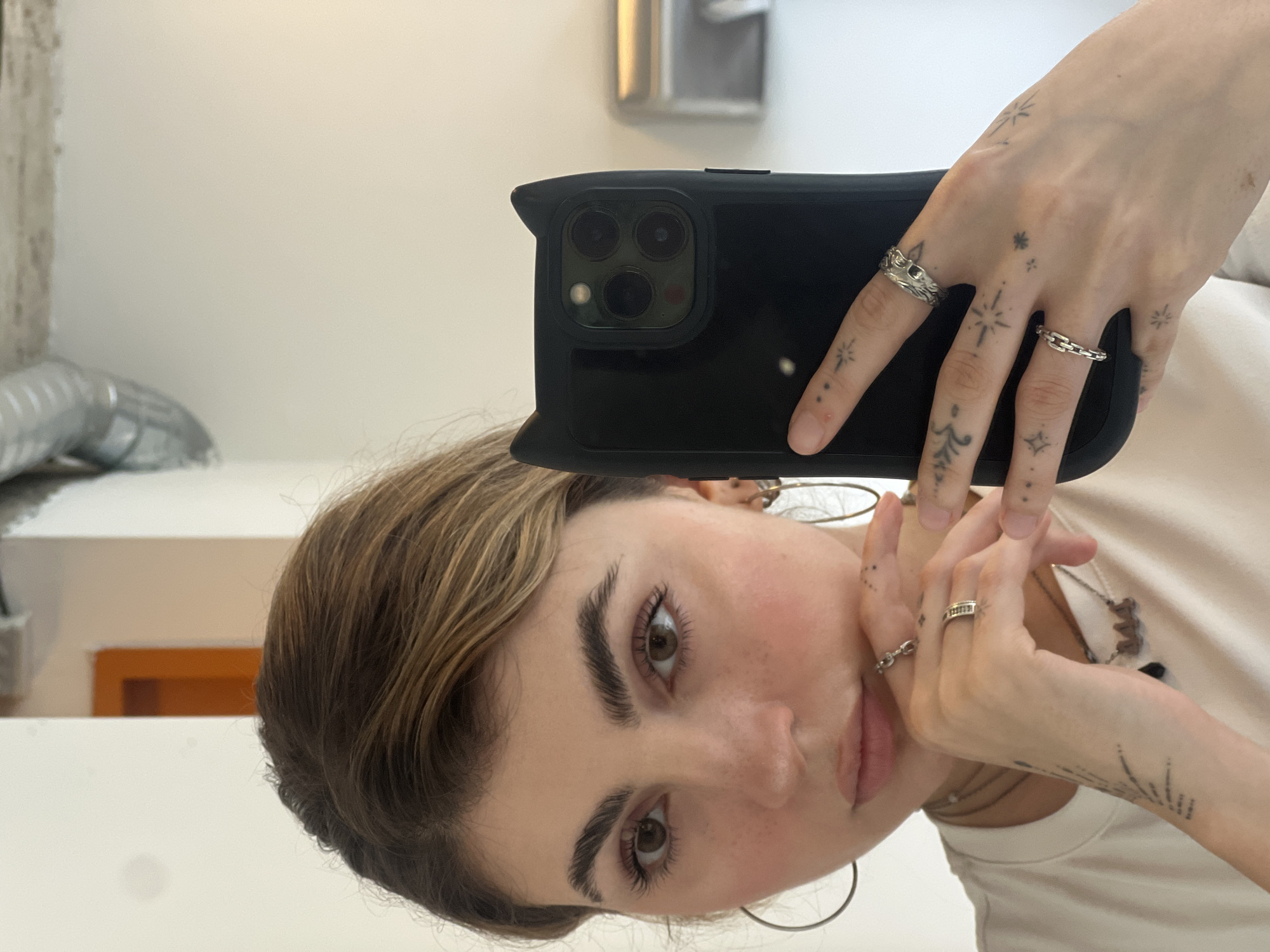A vertically oriented mirror selfie depicts a young person, presumably female, holding a black smartphone with a case to capture their reflection. The subject has light brown hair, prominent dark eyebrows, and brown eyes. Freckles are visible across their nose and cheeks. They wear large hoop earrings and a light-colored t-shirt, along with several necklaces. Their hands are adorned with numerous small, delicate tattoos, featuring stars, dots, and abstract designs. Multiple silver-toned rings, including one with a wave pattern and another resembling a chain link, are worn on their fingers. The smartphone's camera array, featuring three lenses and a flash, is clearly visible. The background is mostly a plain white wall, reflecting an orange rectangular element, possibly a door frame or furniture, and a metallic object in the upper left, suggesting an indoor setting like a home or studio in Sofia, Bulgaria. No text is visible in the image. The lighting is bright, indicating daytime or well-lit artificial light. The subject's expression is neutral and calm.