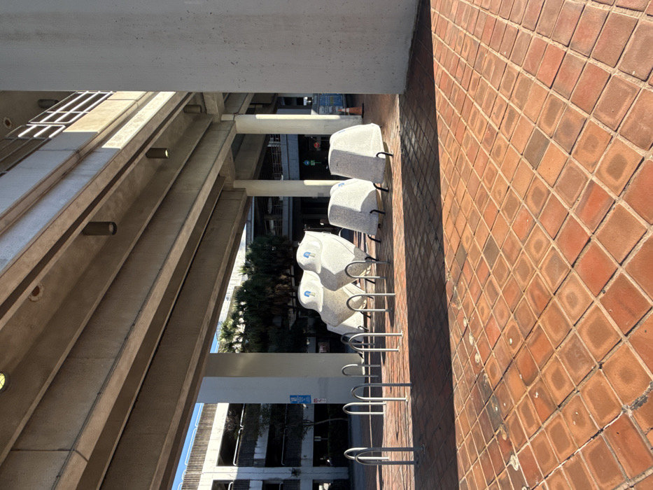 A row of covered stone benches lines a brick walkway under an architectural structure. The benches are white with gray flecks and metal supports. Bike racks are positioned in front of the benches. A multi-story building and trees are visible in the background. The bricks of the walkway are set in a pattern and are various shades of brown.