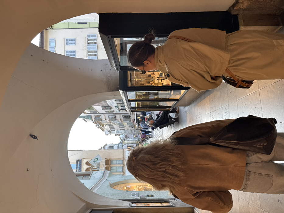 The image depicts an arcaded pedestrian thoroughfare in Bolzano, Italy, likely the Portici (Laubengasse), viewed from beneath the arches towards a bustling street scene.
In the immediate foreground, two female subjects are visible from behind. On the left, a woman with long, wavy blonde hair, secured with a light-colored clip, wears a tan or light brown suede-like jacket and light blue jeans. A dark brown hobo-style bag is slung over her left shoulder. She is positioned as if walking forward, away from the camera. On the right, a second woman with dark brown hair styled in a low bun wears a light beige or tan trench coat, consisting of a cropped jacket over a matching skirt or dress. She carries a small, structured brown handbag with a patterned design, worn cross-body or on her right side, and has sunglasses on her face/head. She appears to be stationary or moving slowly, looking towards the right.
Between the two subjects, a series of illuminated glass display cases with dark frames are visible on the right side of the walkway, showcasing various items. The tiled or paved walkway extends into the distance.
Beyond the immediate arcade, a street scene unfolds. Numerous pedestrians are present, walking and congregating. To the right of the street, an older male individual with short hair or balding is seated on a dark metal bench, facing towards the right. Buildings of varying heights line the street, featuring traditional European architecture with multiple windows, some with light blue shutters. A light-colored building on the left exhibits a green flag and decorative elements. The sky, visible at the end of the arcade, indicates daylight, possibly late afternoon or early evening.