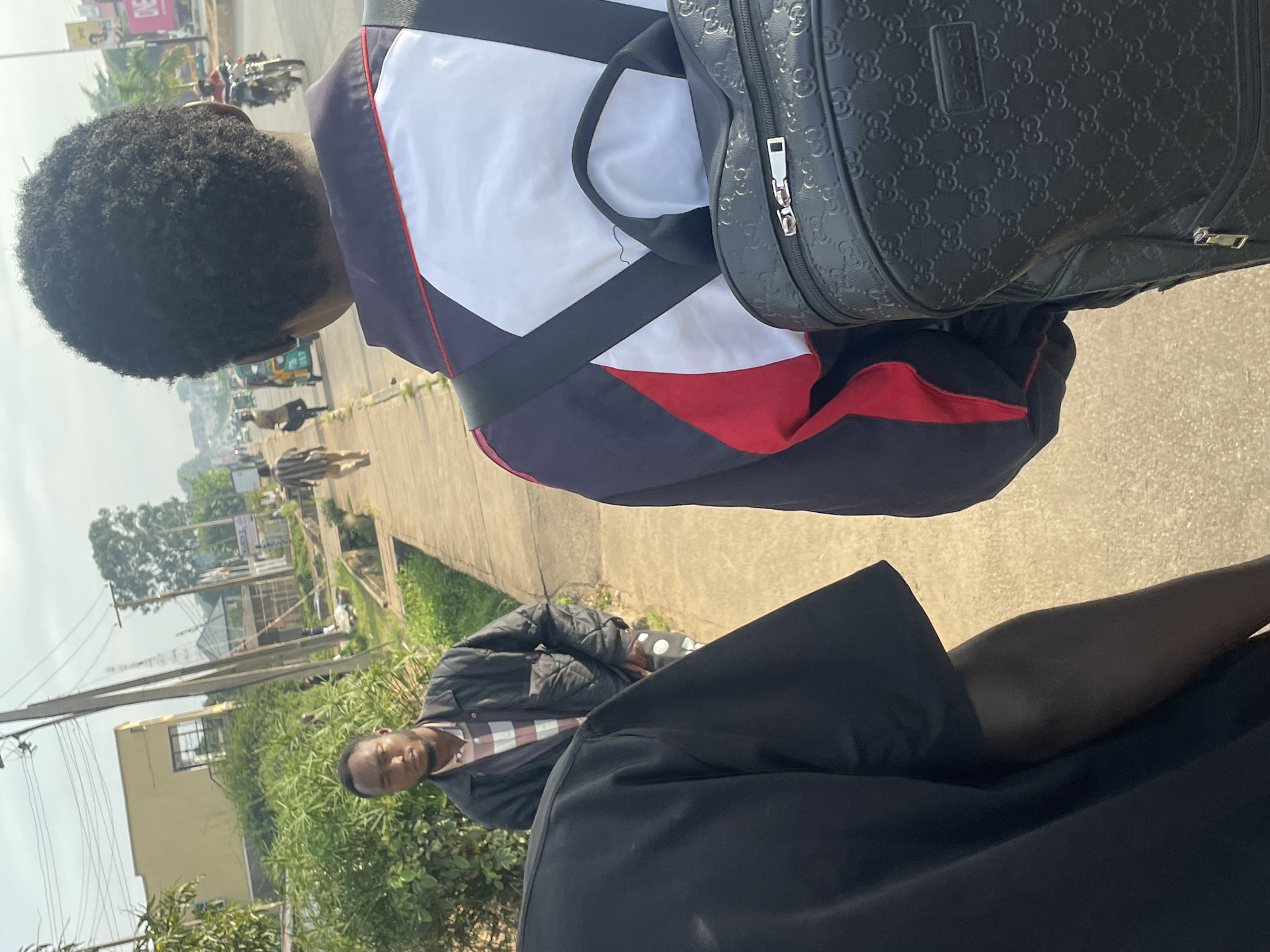 The image depicts a street scene in Jos, Nigeria. In the foreground, two individuals are visible from behind, one wearing a black t-shirt and the other a jacket with white, red, and dark blue panels, carrying a black backpack with a Gucci pattern. To their left, a man in a quilted black jacket and a plaid shirt stands near some greenery. Further down the sidewalk, other individuals can be seen walking, including a person in a striped shirt and another in a yellow shirt. A three-wheeled vehicle is also present on the street, along with a motorcycle carrying a rider. Buildings and utility poles line the street, suggesting an urban environment. The sky is overcast, and the overall lighting indicates it is daytime.