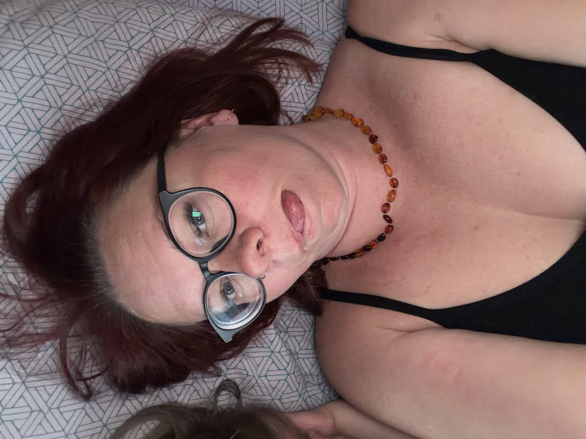 A woman with reddish-brown hair is lying on a patterned bedspread. She is wearing black-rimmed glasses and a black tank top. She also wears a necklace made of amber-colored beads. Her tongue is slightly protruding. A part of a head with brown hair is visible at the bottom of the image.
The image does not provide enough information to determine the location as Chaikovsky, Russia, or any other specific city.