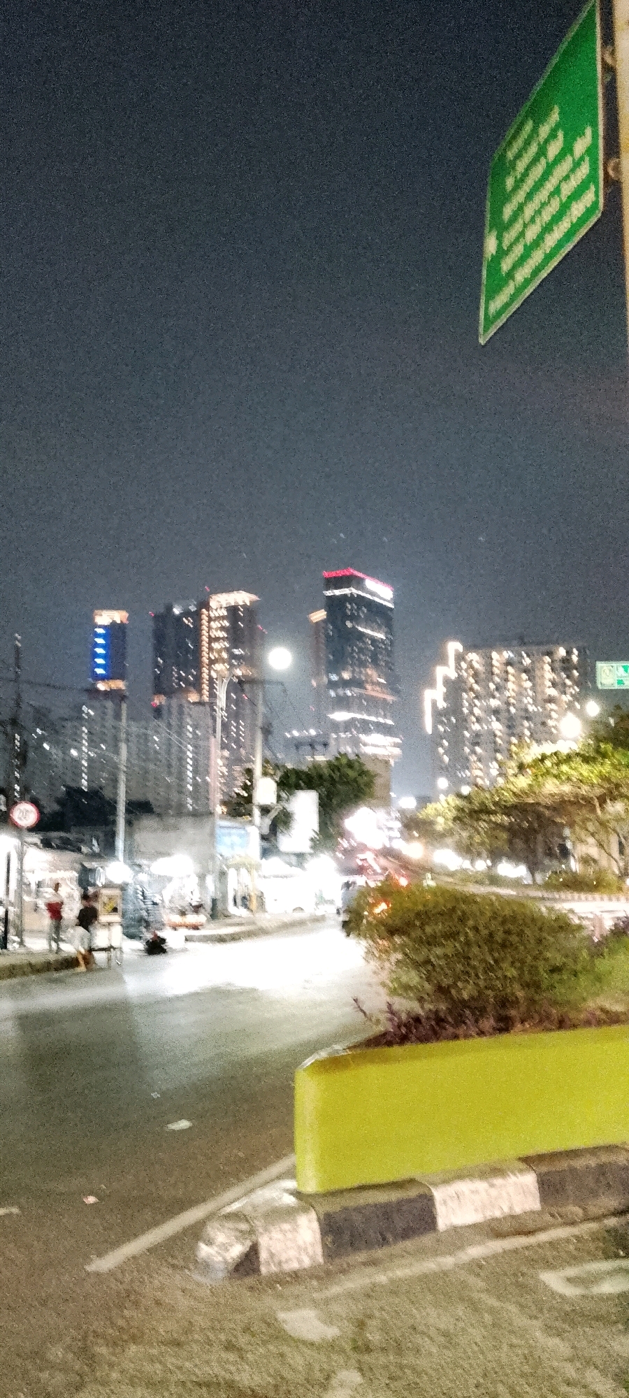 The image captures a nighttime scene on a street in Bekasi, Indonesia, characterized by a mixture of urban elements and pedestrian activity. Several modern high-rise buildings, illuminated by various colored lights, dominate the background, indicating a developed city environment. A street lamp illuminates the road, which appears wet, suggesting recent rain. On the sidewalk, several figures are present, some appearing to be vendors with stalls, while others are pedestrians. The foreground features a bright green barrier and a striped curb, common urban landscaping elements. A green directional sign with white text is visible in the upper right corner. The overall impression is one of a busy urban area at night.