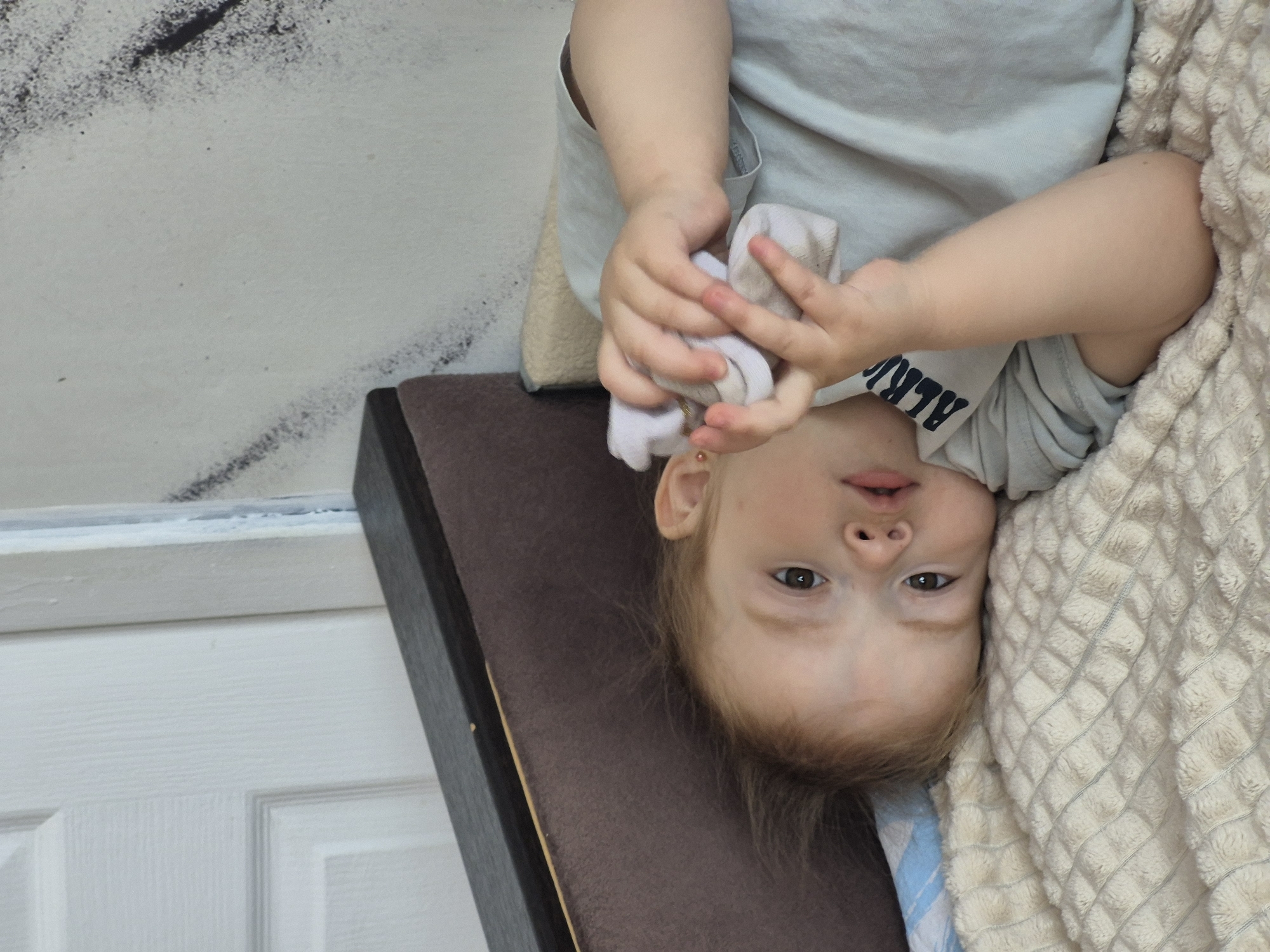 In an indoor setting, a young child with blond hair lies on a textured, cream-colored blanket. The child is wearing a light blue t-shirt with dark lettering that is partially visible. The child's head is tilted, and their right hand is holding a white piece of fabric near their face. The background shows a portion of a white door on the left and a wall with some dark, abstract markings on the right. There is also a dark brown, cushioned headboard or furniture piece visible behind the child. The scene appears to be in a domestic environment, possibly a bedroom, within the city of Chaikovsky, Russia.
