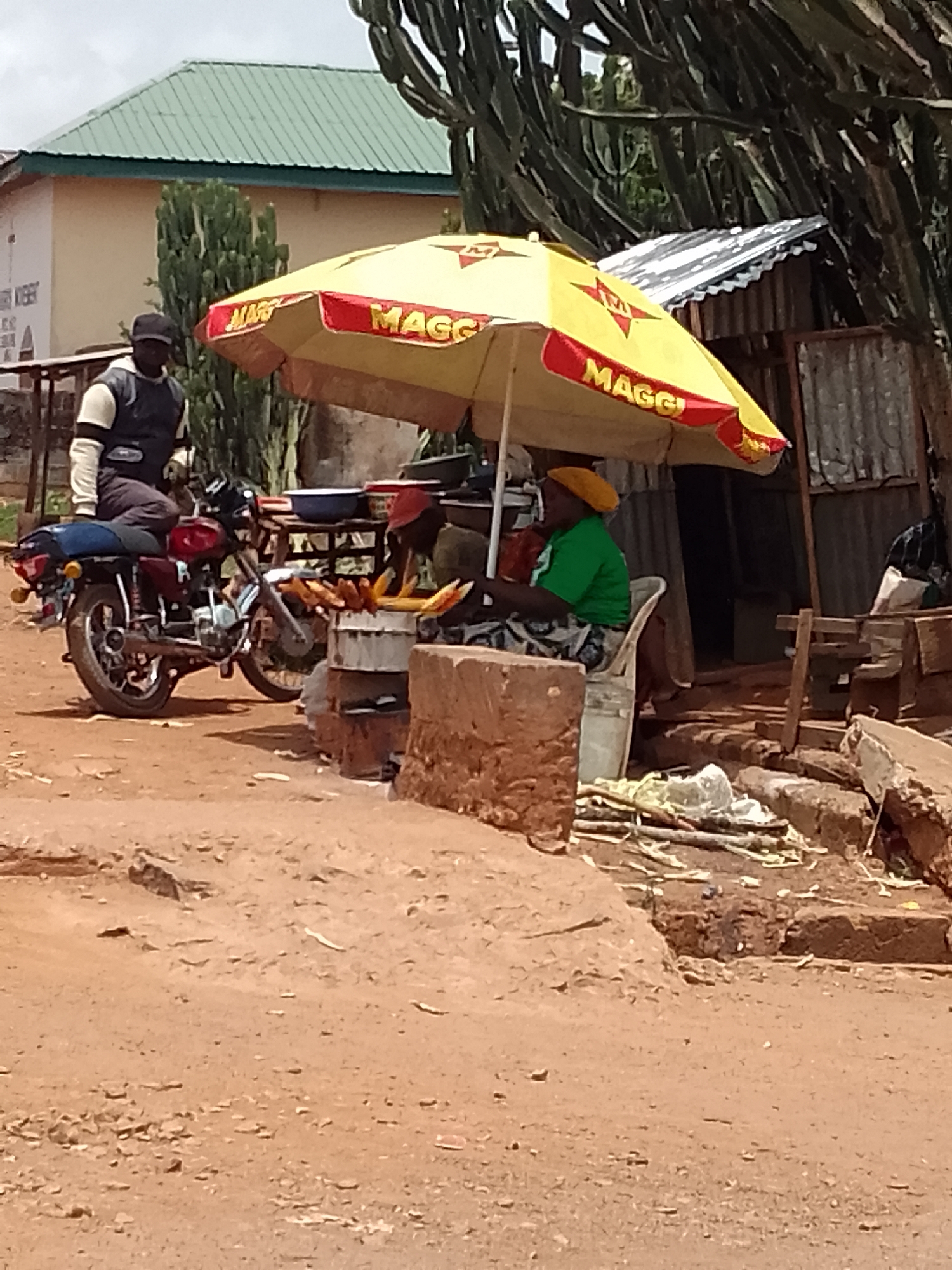 The image depicts a street scene in Vom, Nigeria. A man is sitting on a motorcycle parked on a reddish-brown dirt road. Nearby, under a yellow and red "Maggi" branded umbrella, a woman wearing a green shirt and yellow head covering sits in a white plastic chair. She appears to be tending to a makeshift food stall, where grilled corn on the cob is displayed. The stall is constructed from rough brick or stone. Behind the stall is a corrugated metal structure, possibly a small shack or store. In the background, there is a building with a green roof and some cactus-like plants. Debris and sticks are scattered around the stall area.