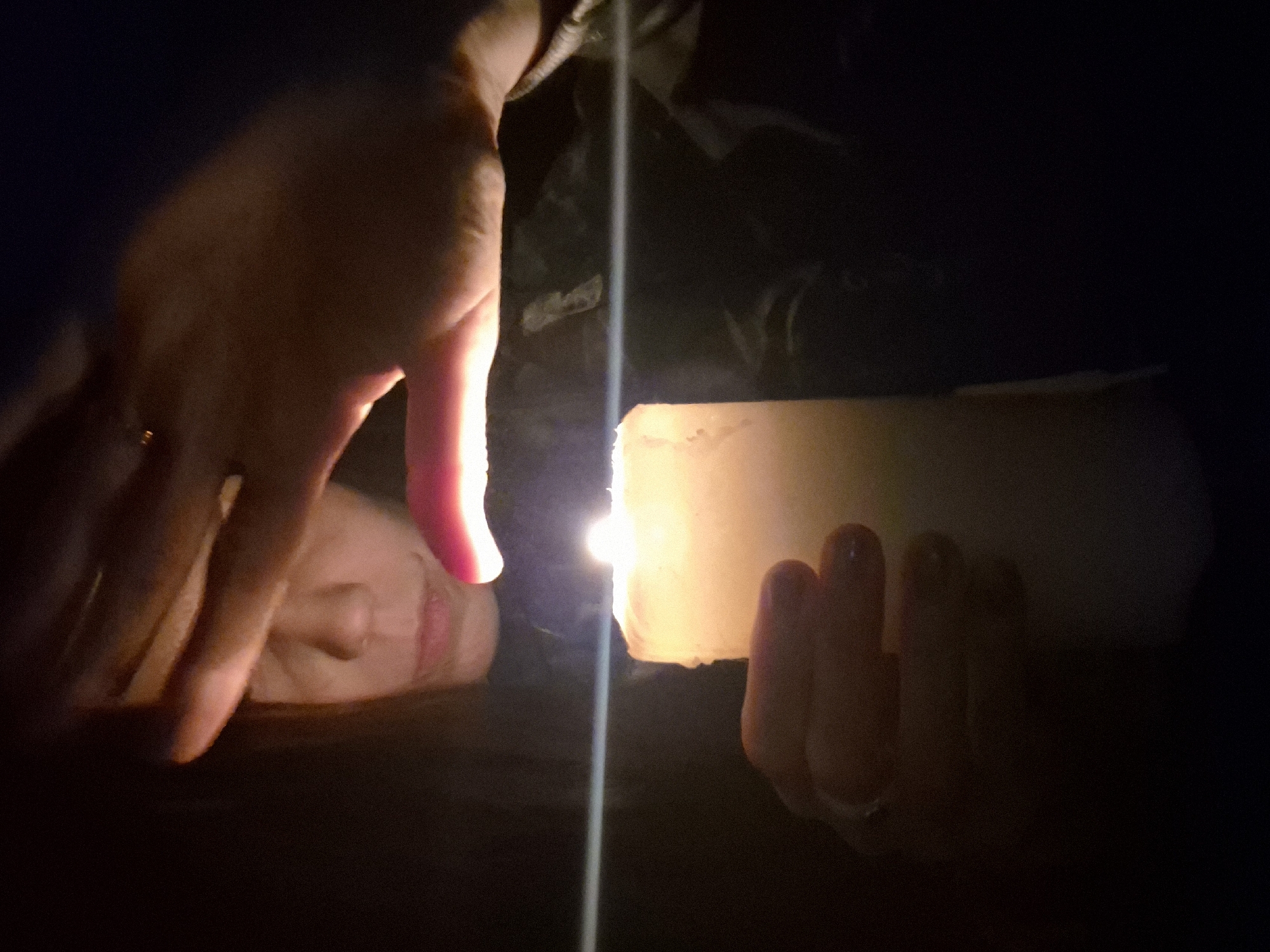 This intimate photograph captures a close-up scene predominantly lit by a single candle in a dark setting, likely indoors in Chaikovsky, Russia. The primary subjects are a person's hands and a thick, white candle.
One hand, adorned with a ring on the index finger, is positioned on the left side of the frame, reaching towards the light. Its fingers are spread, and the hand is bathed in the warm, orange-yellow glow of the candle flame, creating strong highlights and shadows. A second hand, holding the candle from below, is visible on the right, with its fingers also illuminated. The candle itself is substantial, appearing almost cylindrical, and its rough texture is visible under the intense light from its wick.
In the blurred background, partially visible and dimly lit by the candle's glow, is a person's face, turned sideways, suggesting a contemplative or focused posture. The extreme low light conditions indicate that the image was taken at night or in a completely darkened room. A distinct, bright vertical streak of light runs through the center of the image, cutting across the candle flame and extending upwards and downwards, possibly a lens flare or reflection.
The overall mood conveyed is one of quiet contemplation, intimacy, or perhaps a moment of ritual, emphasized by the solitary light source and the focused interaction with the candle. There is no visible text in the image.