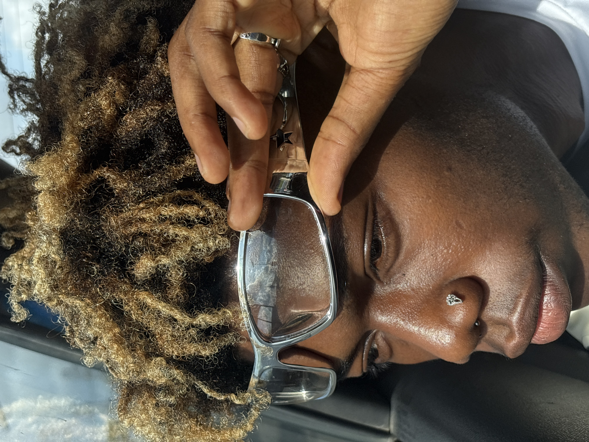 A dark-skinned person with brown and black curly hair is looking at the camera through clear, silver-rimmed glasses. The person has a silver ring on their finger and a silver stud in their nostril. The picture was taken during the day with sunlight and shadow present. Based on the background color, they may be sitting in a car. The location in the city of Ikoyi, Nigeria cannot be determined from this image.