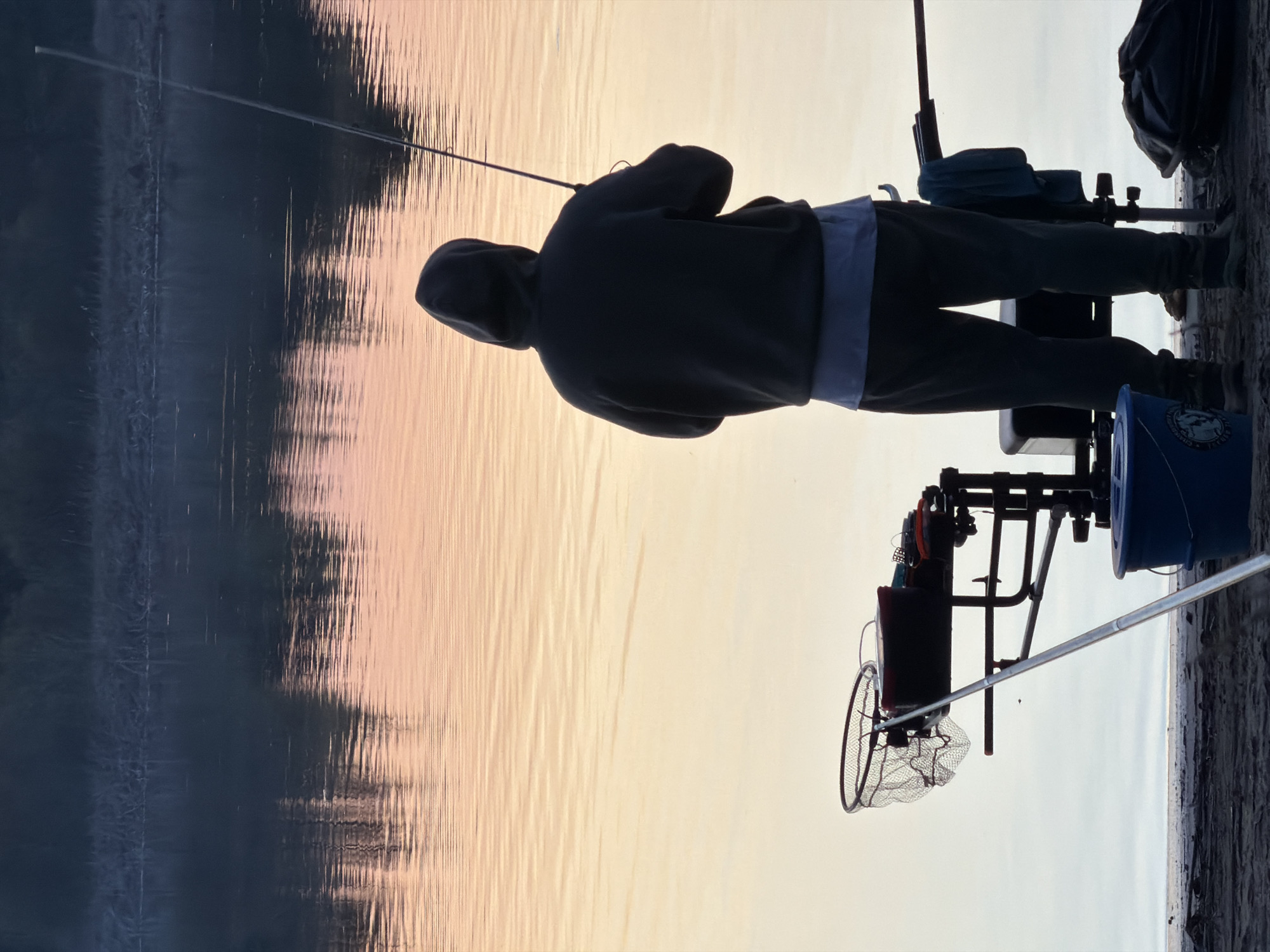 The image shows a person fishing by a body of water, possibly near Chaykovsky, Russia. The person is silhouetted, wearing a hooded sweatshirt, and holding a fishing rod. Fishing equipment such as a net, tackle box, and bucket are visible near the person. The background is split between a dark, reflective body of water on the left and a bright sky or water surface reflecting the sky on the right.