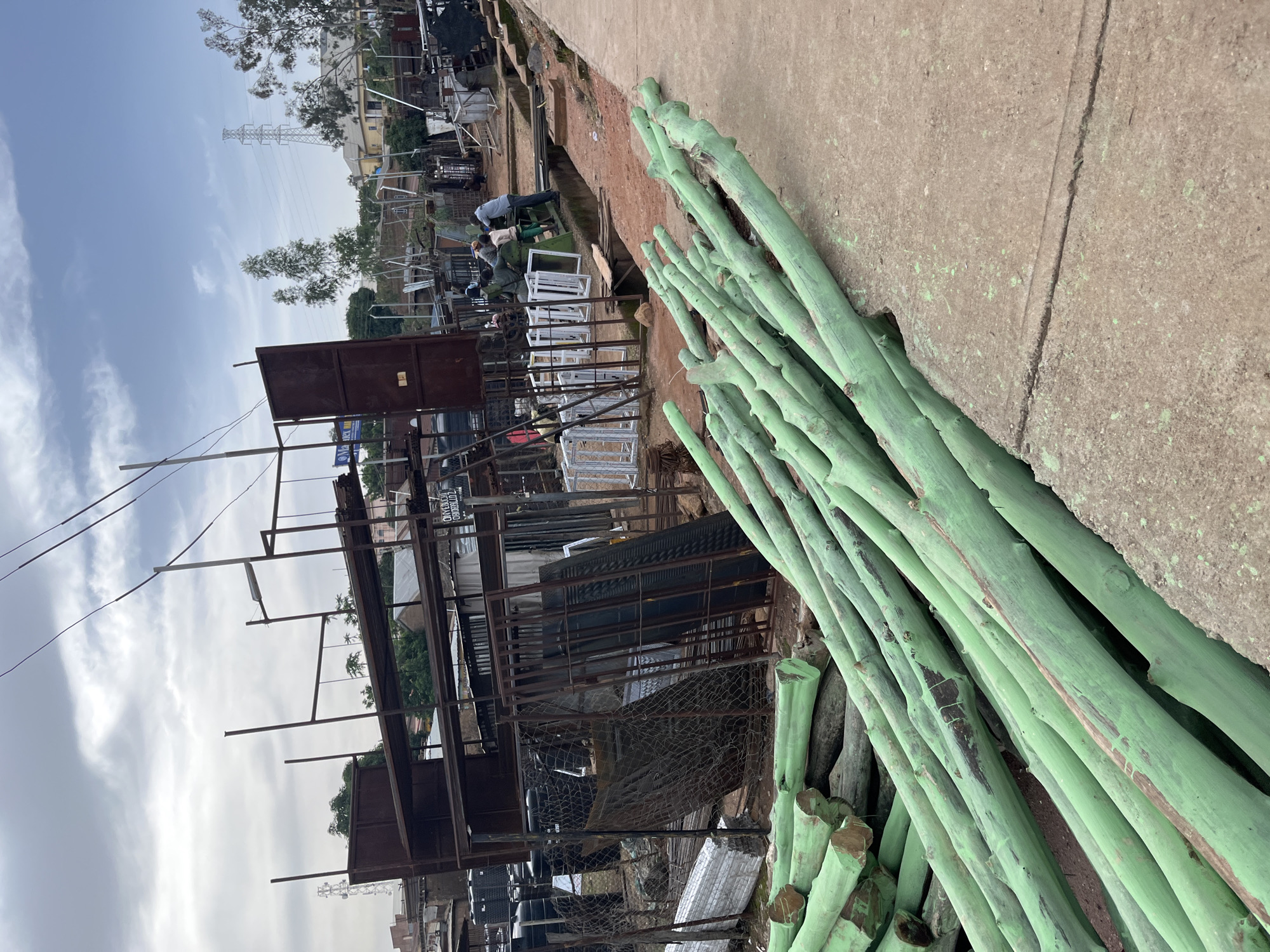The image depicts an outdoor scene, likely a junkyard or a scrap metal processing facility, in Gura Top, Nigeria. A prominent feature is a large pile of thick, green-painted wooden poles or logs lying diagonally in the foreground on a concrete surface. Behind the logs, the scene unfolds to reveal a chaotic arrangement of metal structures, possibly part of the facility. These structures include racks, frames, and corrugated metal sheets, suggesting the storage or processing of salvaged materials.
In the middle ground, several white-framed window units are visible, leaning against a metal structure. Further back, there are more metal objects, including what appear to be black barrels. A few people are subtly visible in the background, engaged in activities that are not clearly discernible but suggest ongoing work. One person is bending over a table, and others are standing or moving around the area.
The overall setting appears industrial or utilitarian. The sky is overcast with a mix of clouds, indicating a daytime scene with possibly dull weather. There are some trees and a distant power line tower, providing a glimpse of the surrounding environment. The mood of the image is practical and work-oriented, focusing on materials and the environment of a salvage yard. No legible text is apparent in the image.