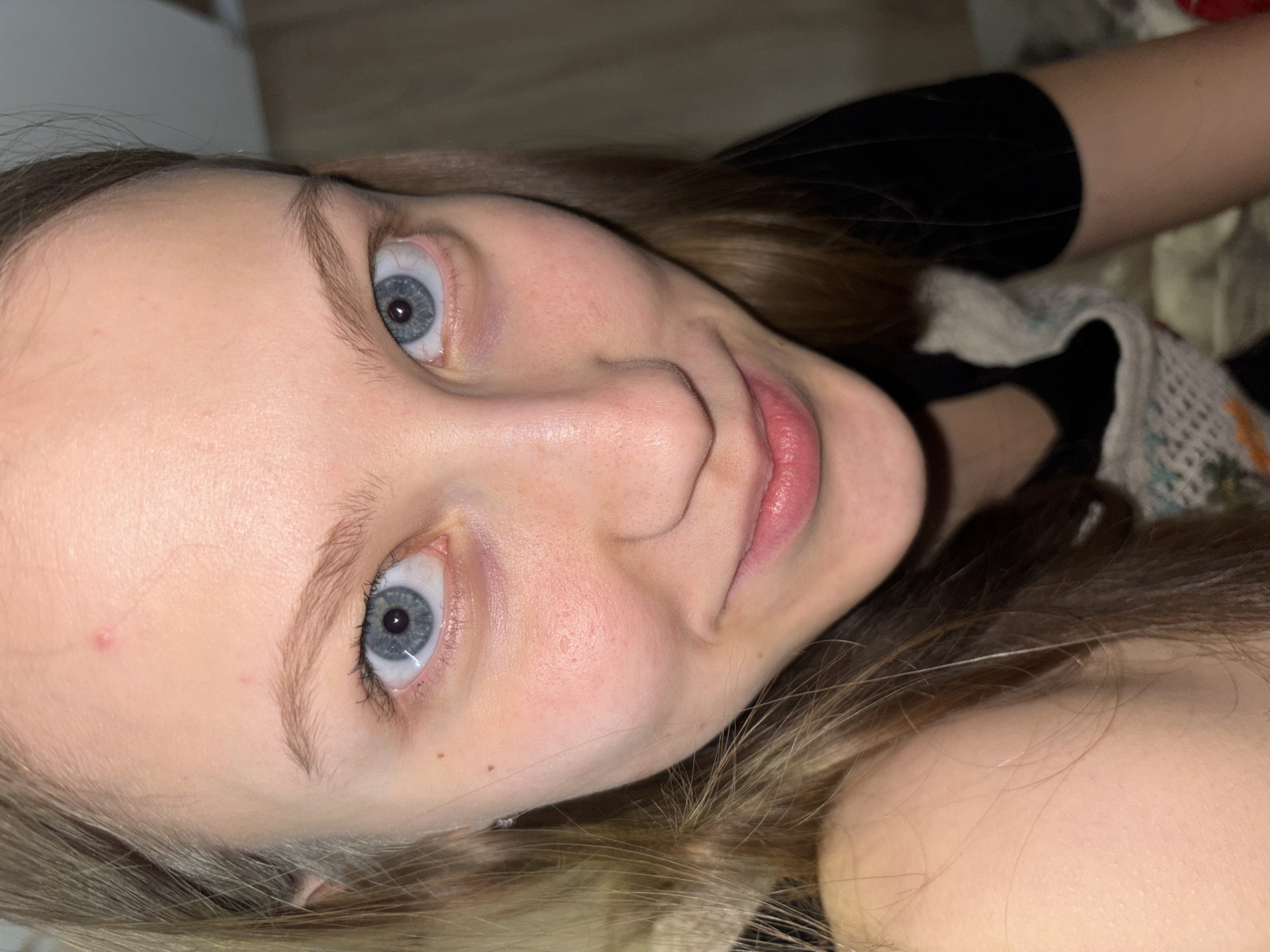 Close-up, angled view of a Caucasian woman's face with blue eyes, light skin, and blonde hair. The background is indistinct but appears to be an interior space with fabrics and dark clothing visible. There's no discernible information to place the scene in Perm, Russia.