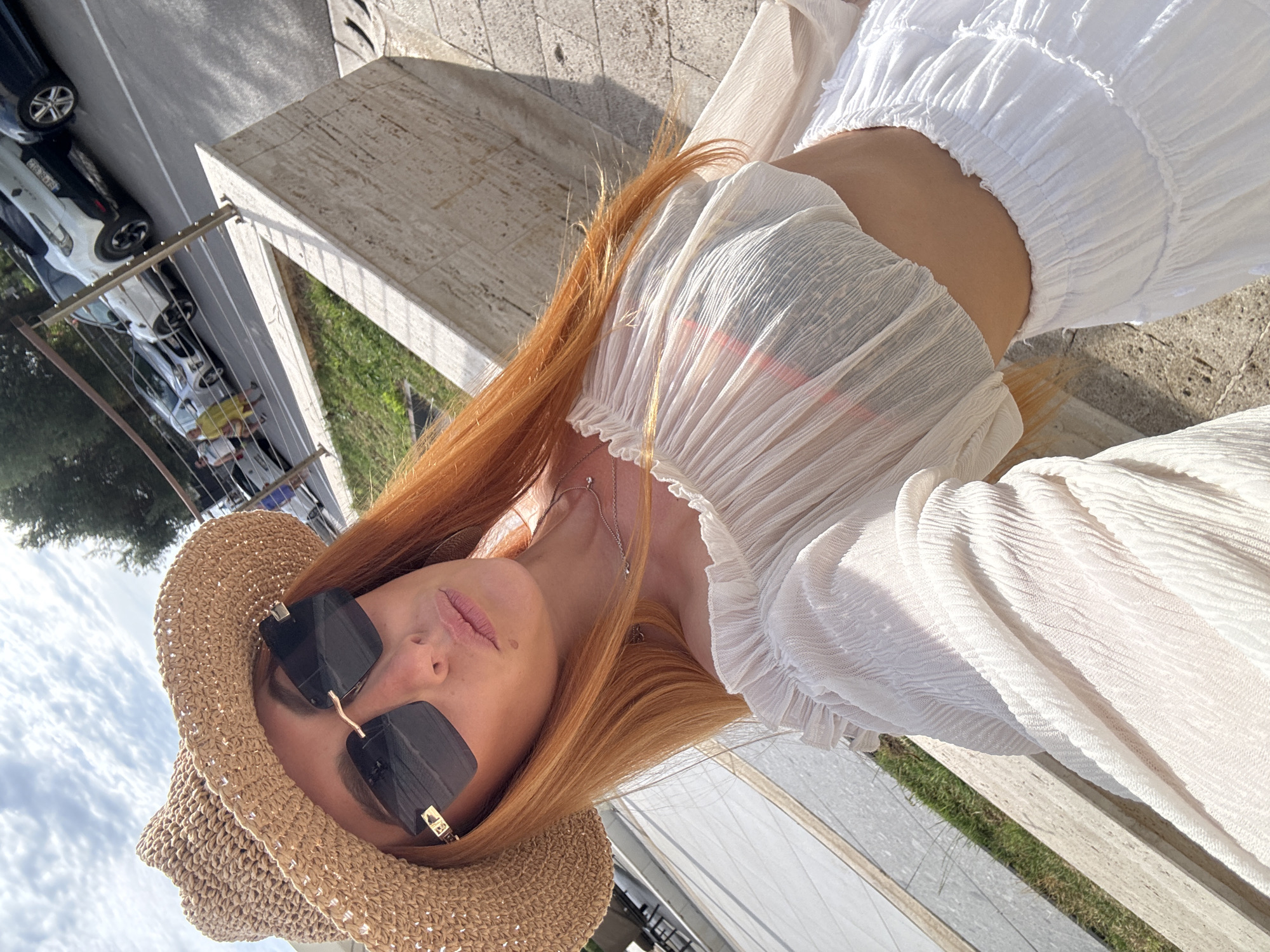 An outdoor, high-angle selfie captures a light-skinned woman with long, reddish-orange hair, wearing a woven straw hat and dark, rectangular, rimless sunglasses with gold-colored accents. She is dressed in an off-white, sheer, textured off-the-shoulder crop top, revealing a dark undergarment beneath, and a white, tiered, elastic-waist skirt, exposing her midriff. A layered silver-colored necklace with small pendants is visible around her neck.
The background depicts a sunny street scene in Petrovac na Moru, Montenegro. A beige concrete wall with a metal railing (vertical posts, horizontal bars) separates the foreground from an asphalt road with white parking lines. Several vehicles are parked along the road, including a white Renault Captur with a visible Montenegrin license plate displaying "BD BG818" and a dark blue/black car. Multiple individuals are walking on the sidewalk beyond the parked cars, one person distinctly wearing yellow. To the left, a patch of green grass is visible beside a light-colored paved area. The sky above is partially cloudy blue.