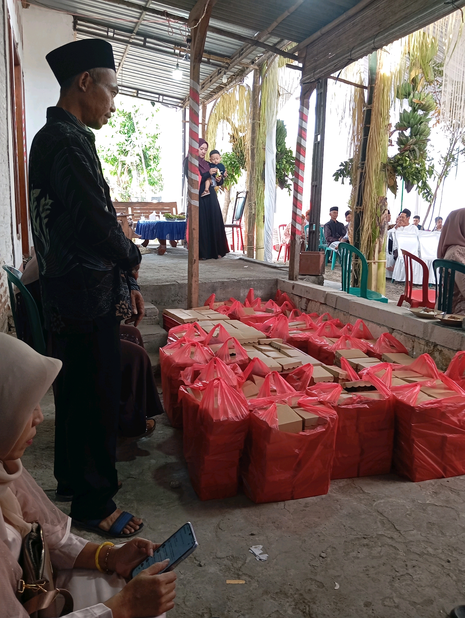 The image depicts an outdoor event with a contingent of people gathered under a covered structure. In the foreground, to the left, an elderly man wearing a black peci and a black patterned shirt stands in profile, facing right. To his right, a woman wearing a light-colored hijab and a light-colored outfit is seated and looking down at a smartphone in her hands. Behind them, a large number of red plastic bags containing brown boxes are arranged on the ground, suggesting prepared meals or gifts.
In the background, several individuals are seated on chairs, some wearing traditional Indonesian attire. A woman in a dark dress holds a baby. The environment appears to be a village setting, possibly in Ngampel, Indonesia, with elements of natural foliage and a rustic roof structure overhead. The overall scene conveys a sense of community gathering and shared provisions.