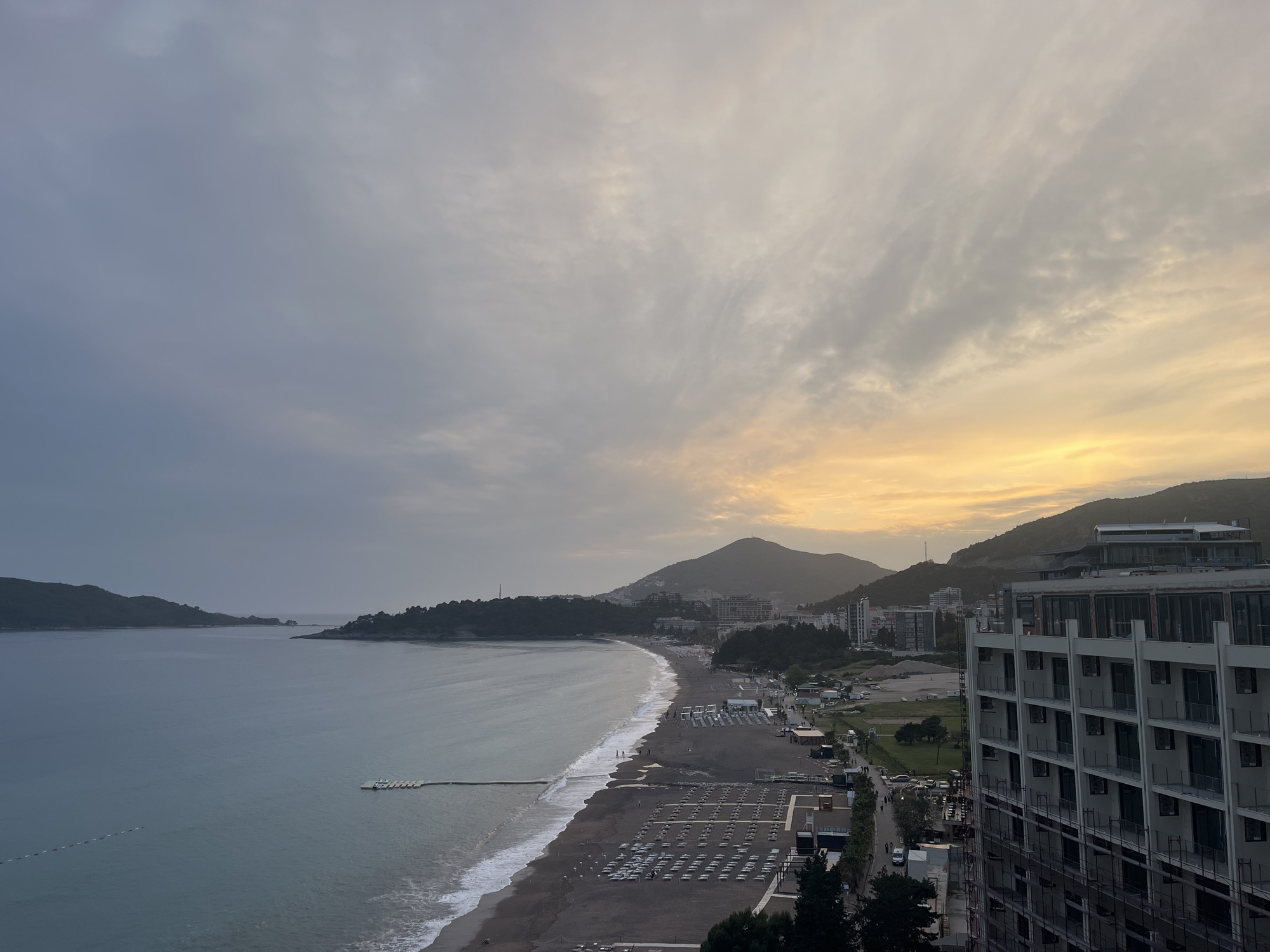 The image shows a coastal view in Rafailovići, Montenegro. The scene includes a beach with dark sand, rows of beach chairs, and gentle waves meeting the shore. In the distance, there's a peninsula and a mountainous backdrop with some buildings scattered along the base of the mountains. The sky is overcast with shades of grey and a hint of golden light towards the horizon, suggesting either sunrise or sunset. A tall modern building is visible on the right side of the frame, partially obscuring some of the background details. People can be seen walking along the promenade adjacent to the beach.