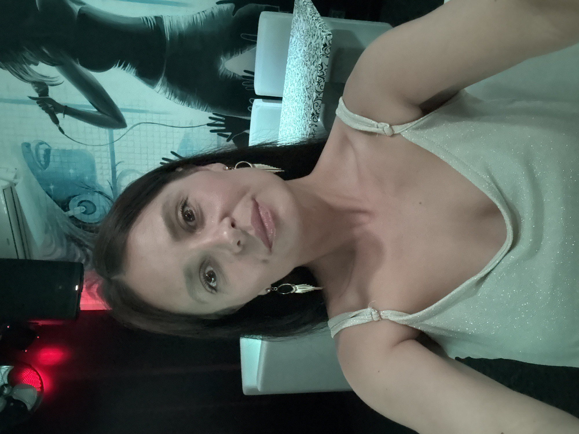 A woman is in a room with a wall mural. The woman has dark hair and is wearing a light-colored dress with thin straps. She is wearing earrings. The wall mural behind her depicts a woman singing into a microphone. The location is in the city of Chaikovsky, Russia.