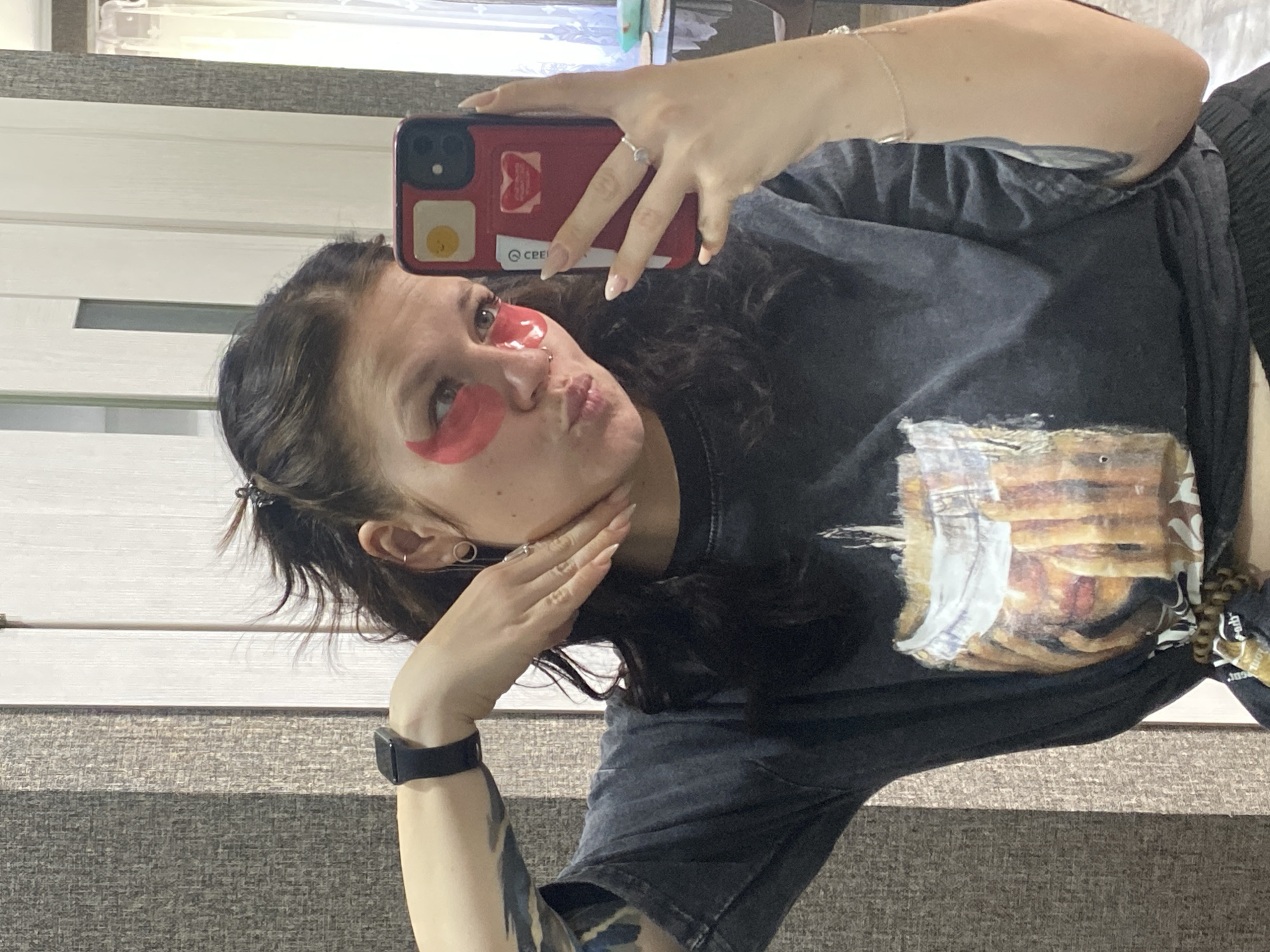 A woman is taking a selfie, with red eye patches under each eye. She is pursing her lips slightly. The woman is holding a red iPhone with stickers, and her other hand is touching her chin. She is wearing a black t-shirt and a black smart watch. Part of a tattoo is visible on her arm. The background includes a gray wall. There is no indication of the city of Chaikovsky, Russia.