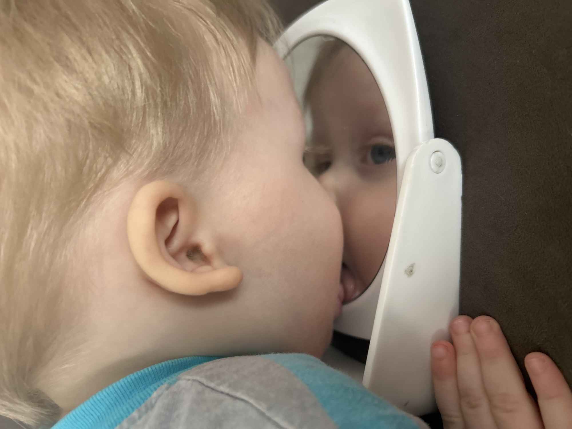 A young child with light blonde hair and fair skin is depicted leaning over and interacting with a white, hinged compact mirror. The child's face is positioned directly above the mirror, with the reflection of their blue eye visible within the glass. The child's mouth or lips appear to be touching or in close proximity to the mirror's surface. A grey garment with a light blue ribbed cuff is visible on the child's left side. A small hand, likely belonging to the child, rests in the lower-left foreground on a dark brown, textured surface. The white plastic frame of the mirror shows a small dark smudge. The background is out of focus and dark. The specific location within Chaikovsky, Russia, cannot be determined from the visual information provided.