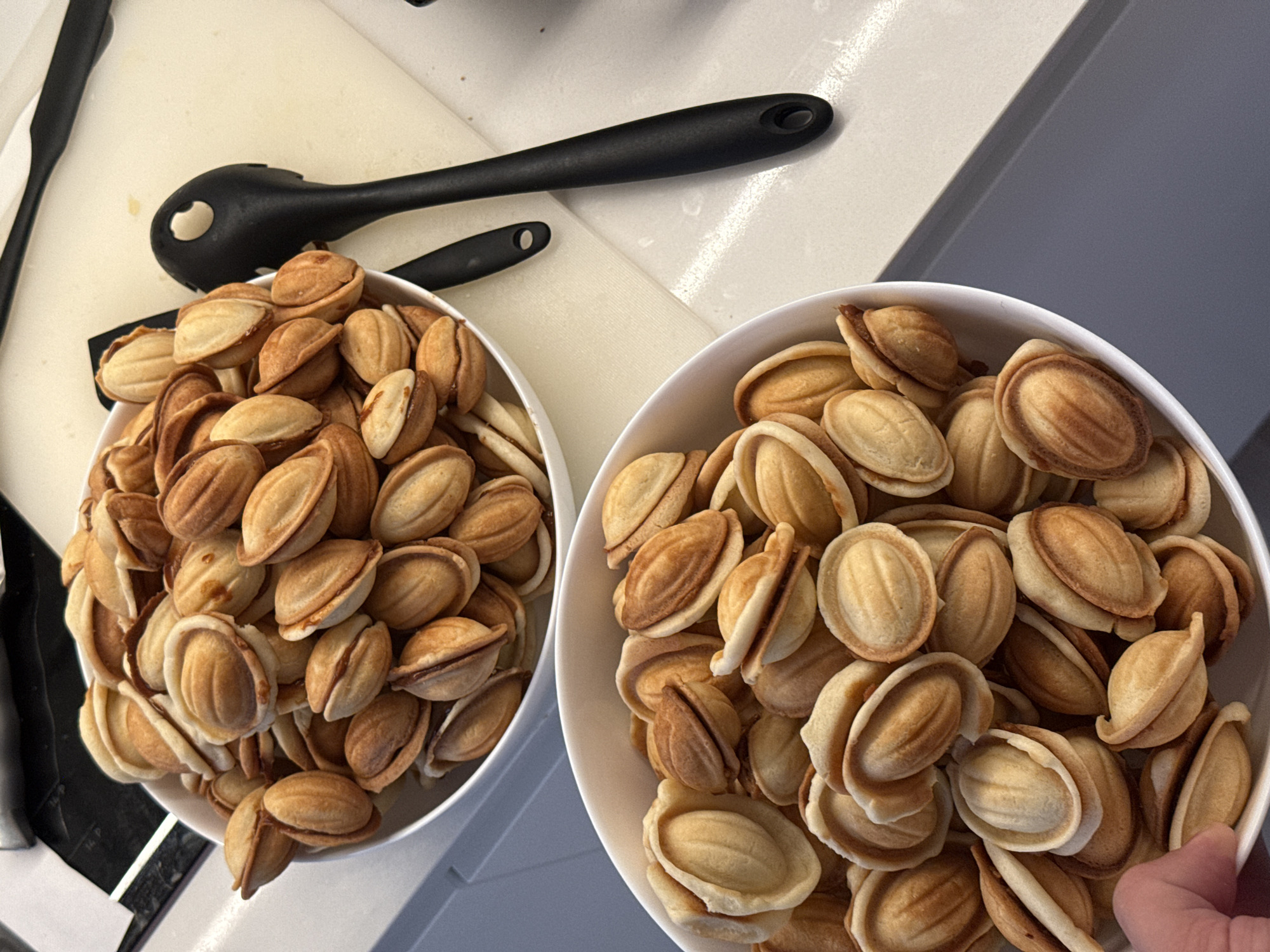 Two white ceramic bowls, each filled with numerous freshly baked Oreshki cookies, are positioned on a kitchen counter in Мокино, Russia. The Oreshki are golden-brown and oval-shaped, resembling small nuts, with visible seams from their molding. The upper bowl rests on a rectangular, light-beige cutting board or mat. Adjacent to this bowl, on the cutting board, are two black plastic kitchen utensils: a spatula and a ladle/spoon, both featuring a circular cutout at the end of their handles. The lower bowl, closer to the viewer, is partially supported on its left side by a human hand, revealing a thumb and multiple fingers with fair skin. The counter surface in the foreground is light gray, while a darker, reflective surface occupies the upper left background. The overall setting is an indoor kitchen with bright, overhead illumination.