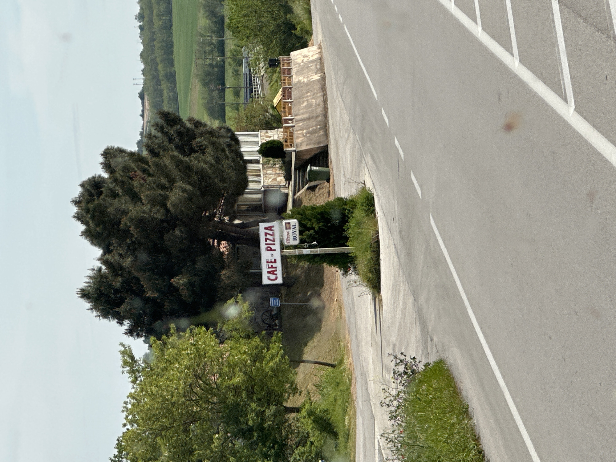The image shows a roadside scene, likely in a rural area of Λύρα, Greece. A cafe/pizza restaurant is situated near the road, indicated by a sign reading "Cafe Pizza". The building appears to be a multi-story structure with a stone facade and balconies. A large, dark green tree dominates the foreground, partially obscuring the view of the building. The road is paved and features white lane markings. Green vegetation, including trees and shrubs, lines the roadside. The background shows a landscape of rolling hills and fields.