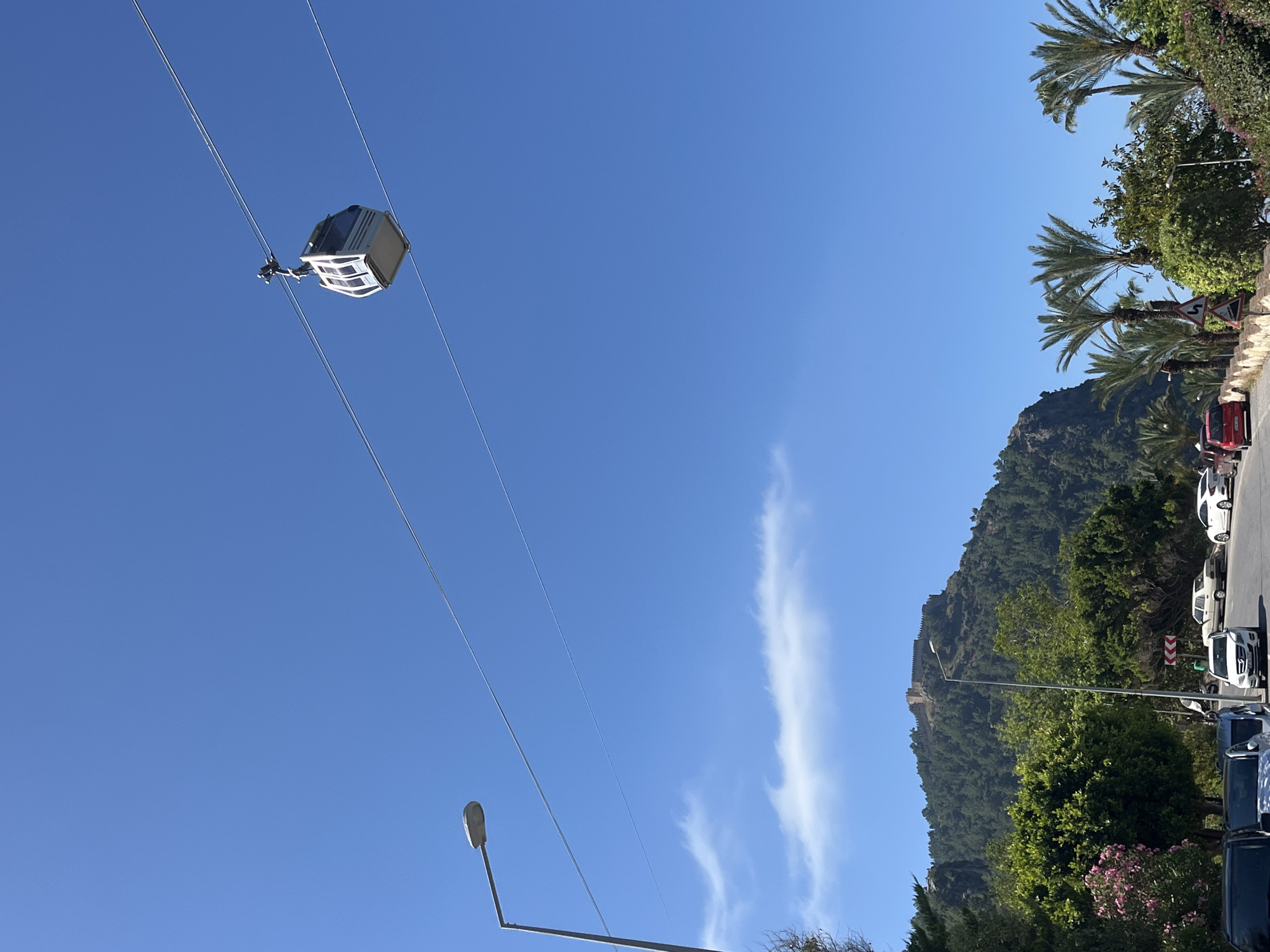 The image shows a cable car ascending against a clear blue sky with a wispy cloud formation. Palm trees are visible in the upper right corner, suggesting a tropical or subtropical environment. In the lower right quadrant, a mountainside covered in greenery is partially visible, with what appears to be a fortress or castle perched on top. Cars are parked along the roadside. The location is Alanya, Turkey.