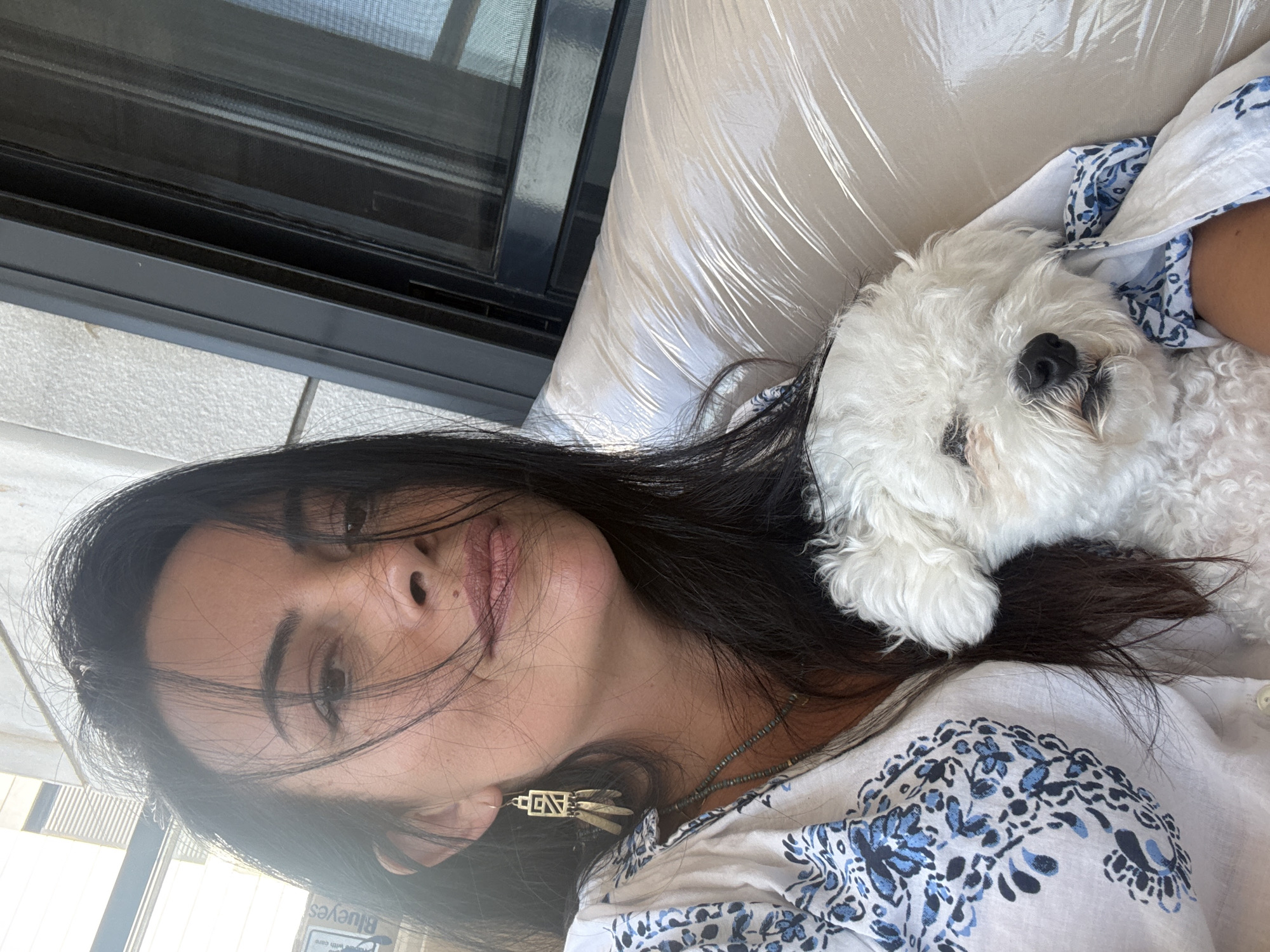 A woman with long, dark hair and a white shirt with blue patterns is taking a selfie outdoors, holding a small white dog. The woman is wearing earrings and a necklace. The dog is nestled in her arms, partially covered by a white cloth with blue designs. Behind them, a wall with horizontal slats is visible, along with a dark-framed window. The setting appears to be a balcony or patio.