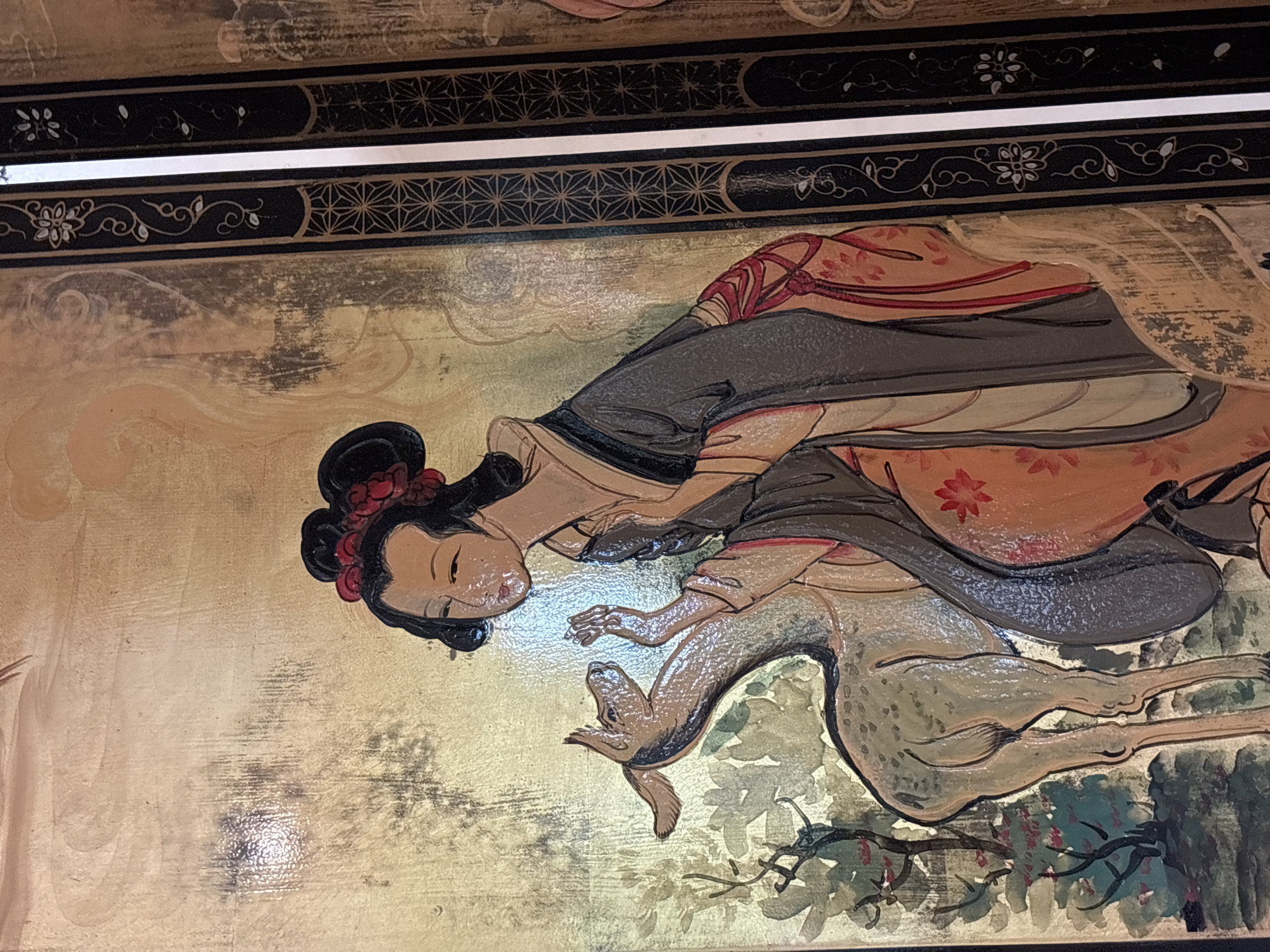 The image is a close-up of an Asian-themed painted screen, featuring a woman and a deer in a garden setting. The woman, depicted in traditional attire with her hair elaborately styled, appears to be interacting with the deer, her hands raised towards its head. The deer is light brown with darker spots. The background is a muted gold with areas of darker shading and hints of green foliage and red flowers. The right side of the image shows a black border with decorative patterns in gold and white. This artwork does not appear to be located in Chalong, Thailand, as there are no contextual elements to suggest a specific geographic location.