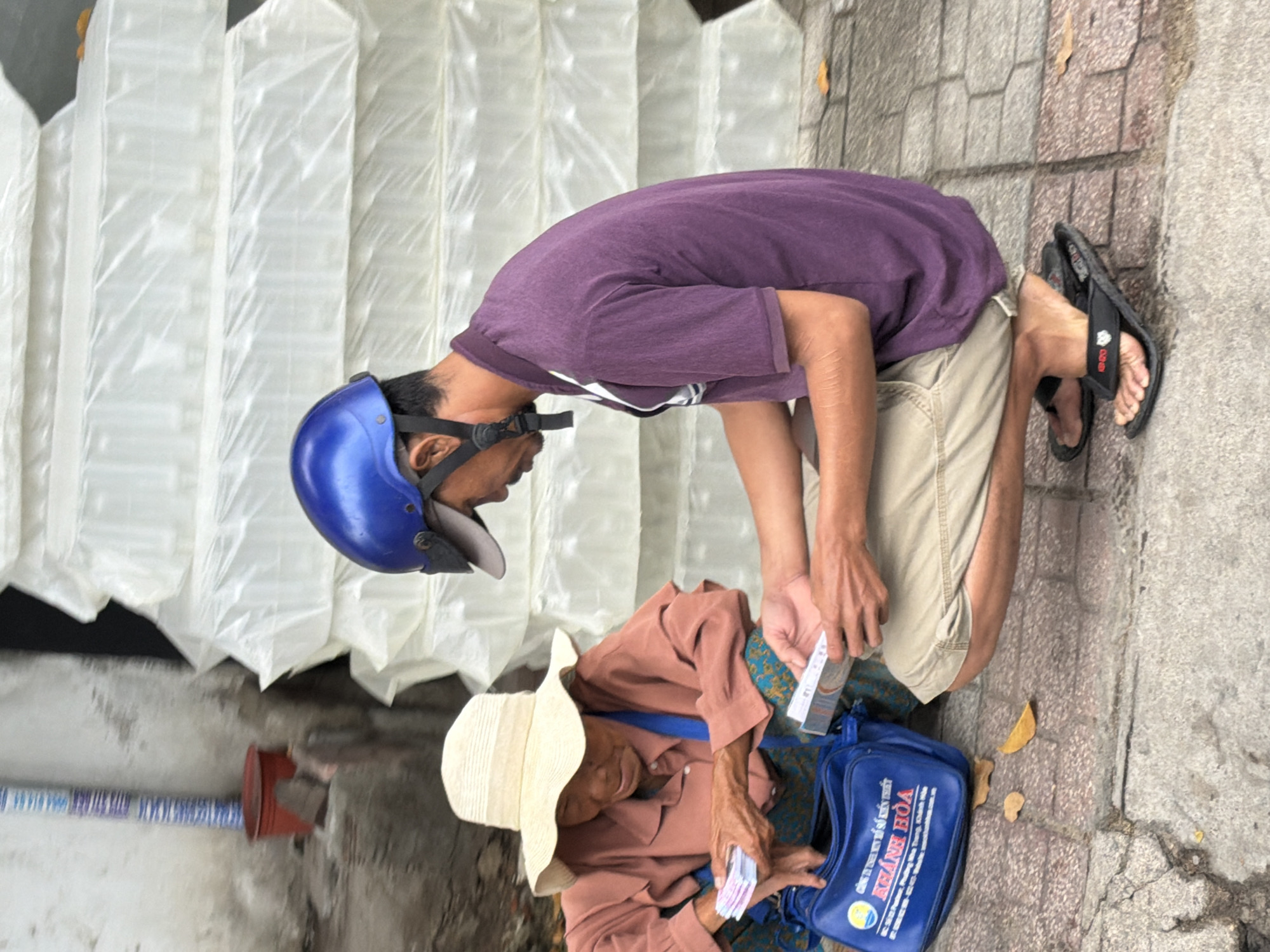 An elderly Vietnamese woman wearing a straw hat and a salmon-colored shirt crouches on a sidewalk in Nha Trang, Vietnam, holding a fan of currency in her hands. A younger man in a purple polo shirt and beige shorts, wearing a blue helmet, squats opposite her, holding a small booklet. In the foreground, a blue bag with Vietnamese text is visible, and a stack of white, plastic-wrapped items is behind the man. The ground is paved with stone and concrete. The scene appears to depict a transaction or exchange between the two individuals on a street in Nha Trang.