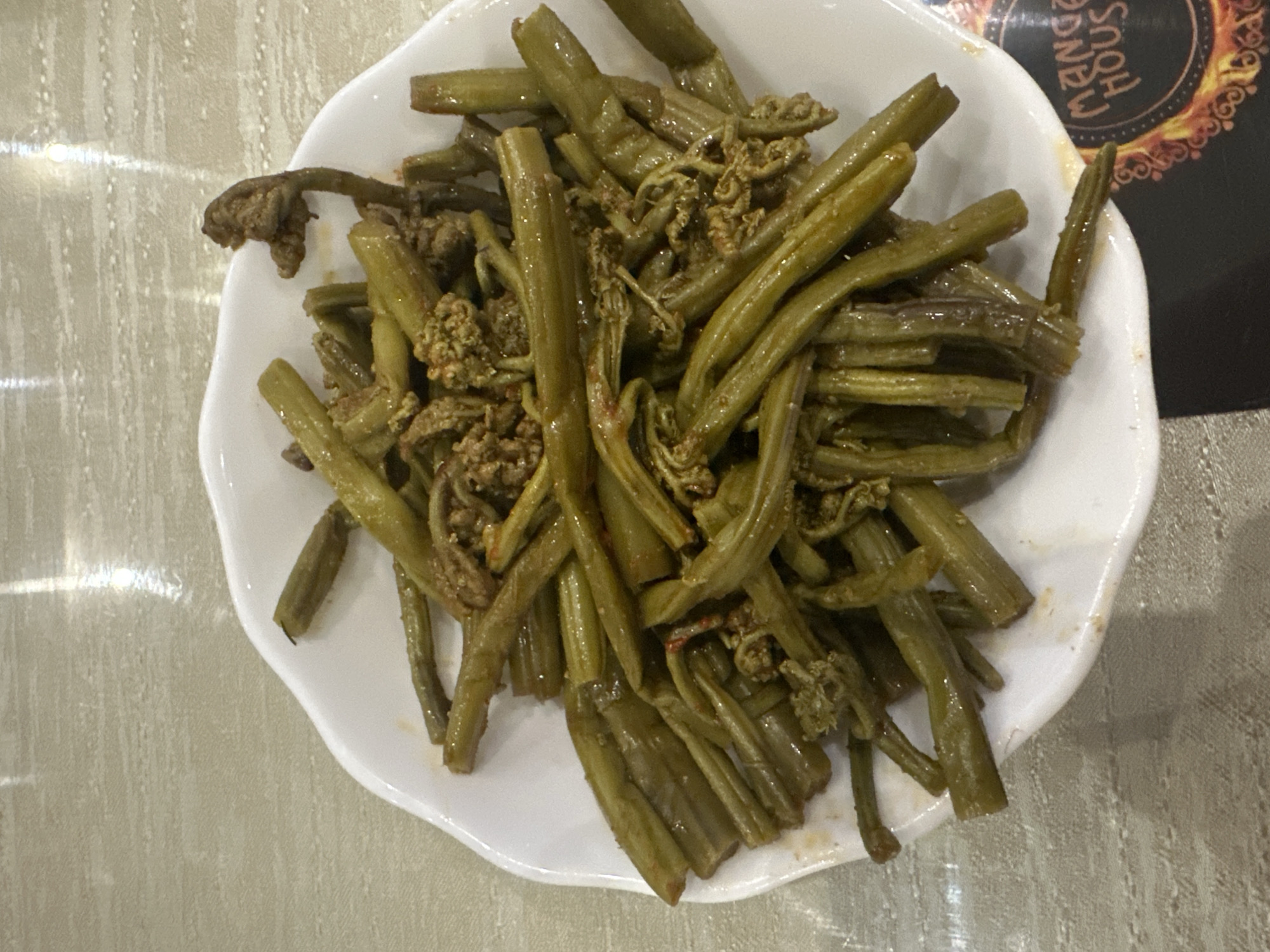 A white, shallow bowl filled with cooked fiddleheads is placed on a light-colored, textured surface. The fiddleheads are green, thick, and appear to be seasoned with a reddish-brown spice mix. Some of the fiddleheads are curled, resembling the characteristic fiddlehead shape, while others are straighter and appear to be cut into segments. The surface beneath the bowl has a subtle pattern and reflects light, suggesting it might be a table. In the bottom right corner of the image, a black coaster with a circular design and the text "MANGO HOUS" is partially visible. No people are present in the image. The scene is shot from a high-angle, looking down at the food. The lighting is bright, casting minimal shadows. The context of Nagornyi, Russia is not discernible from the visual information provided in the image.