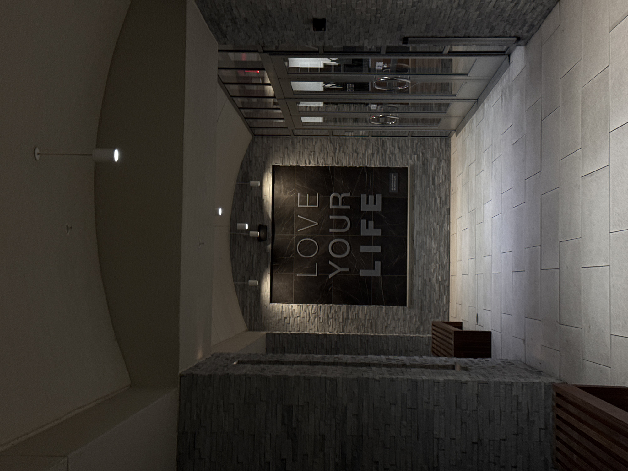 Here is a detailed description of the image:
**Scene:**
The image depicts an interior architectural space, possibly a lobby or entrance hall in a building. The space appears to be in Irvine, California (United States), considering the location specification. The dominant materials are stone or tile, with varying textures and colors.
**Architectural elements:**
- The walls on the left exhibit a curved, arched shape, while the walls on the right are straight and tiled.
- The flooring and some wall surfaces are constructed of a stone or tile brick-like pattern.
- The ceiling is a grid-like framework, possibly a skylight or suspended ceiling with transparent or reflective panels.
- The lighting scheme is a mix of ambient and directed lighting. Spotlights are mounted on the curved wall, and there is likely hidden lighting within the ceiling grid.
**Objects:**
- A large, square, dark-colored plaque with white lettering is mounted on one of the walls, displaying the text "LOVE YOUR LIFE" in a vertical arrangement.
- There are decorative stone or tile structures in the foreground, resembling a low wall or planter box.
- Wooden benches or planter boxes are placed along the tiled wall.
**People and Interactions:**
- There are no visible people in the image. Therefore, there are no interactions or actions to describe.
**Environment:**
- The scene is well-lit, with a mix of natural and artificial lighting. The architectural design suggests a modern and sophisticated style.
- The overall impression is one of a clean, structured, and aesthetically pleasing environment.