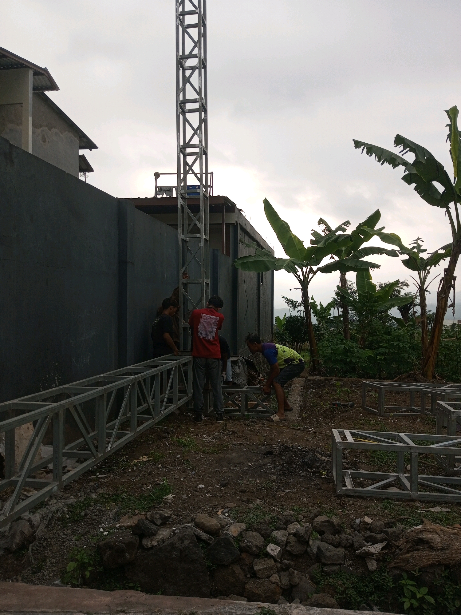 A group of men are working on a metal structure in Andongsili, Indonesia. The scene appears to be an outdoor construction site with a tall metal tower in the background, likely a communication tower. In the foreground, several men are interacting with and assembling metal truss components and cube-shaped frames. One man in a red shirt and jeans is standing near the tower, looking at the structure. Another man in a green and blue shirt is bending over, working on a metal frame on the ground. Two other men are visible behind the man in the red shirt, also seemingly involved in the construction. The ground is covered with dirt, rocks, and some green vegetation. Banana trees and other foliage are present in the background, suggesting a tropical or subtropical environment. The sky is overcast, indicating a cloudy day. A dark, textured wall runs along the left side of the image, with a building visible behind it.