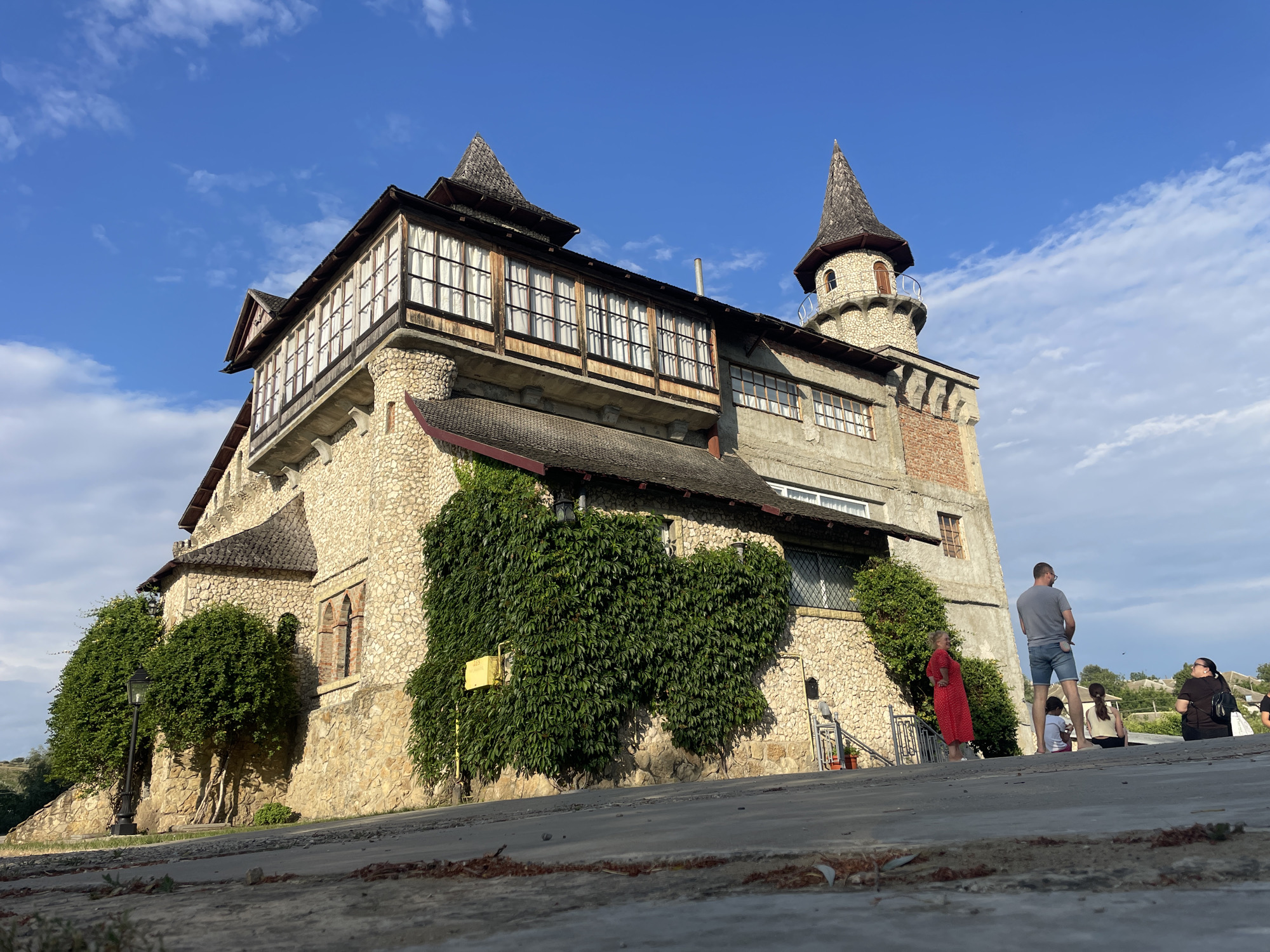 The image depicts a stone-built structure resembling a castle or tower, situated in Colibași, Moldova. The building features multiple levels, a prominent conical turret, and a long, glass-enclosed balcony. The walls are constructed of rough-hewn stones, with some sections covered in dense green ivy. The roof appears to be tiled with a dark material. Several windows are visible, some with visible panes and others in arched openings. The building is set against a backdrop of a bright blue sky with scattered white clouds.
In the foreground, a paved area or walkway is visible, with some debris scattered on it. To the right of the building, a group of people are present. One man, wearing a grey t-shirt and denim shorts, stands facing away from the camera. A woman in a red dress stands near a staircase. Further back, several other individuals are seated or standing, seemingly observing the building or the surroundings. The overall scene suggests a tourist or recreational area.