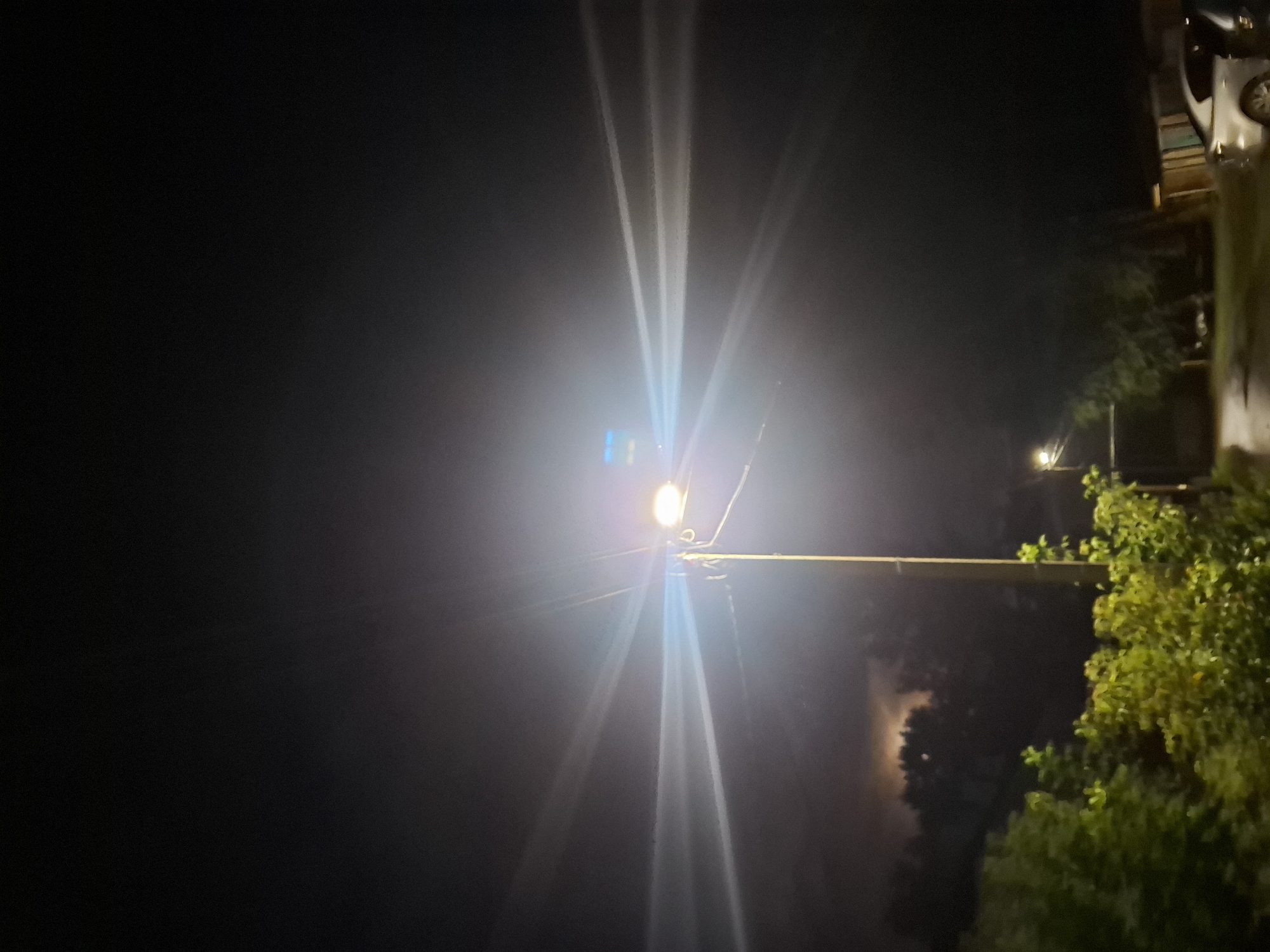 The image depicts a nighttime street scene with a bright streetlamp dominating the center. The streetlamp is tall and metallic, with a single light source emitting a strong, halo-like glow that creates visible light rays fanning out into the darkness. There are power lines attached to the pole. In the background, the sky is dark and overcast, with a faint, hazy illumination suggesting either moonlight through clouds or distant artificial light. Below the streetlamp, trees are visible, some with green foliage, illuminated by the streetlight. To the left, a patch of sky shows a more distinct, diffuse light source, possibly a moon obscured by clouds. On the right side of the image, a residential area is suggested with the outlines of buildings and two parked cars. One car, a silver sedan, is partially visible. The ground appears to be a paved surface, possibly a road or driveway, and there are hints of vegetation along the edges. The scene appears to be an urban or suburban residential area in the city of Novlyanka, Russia. There are no people or animals visible in the image.