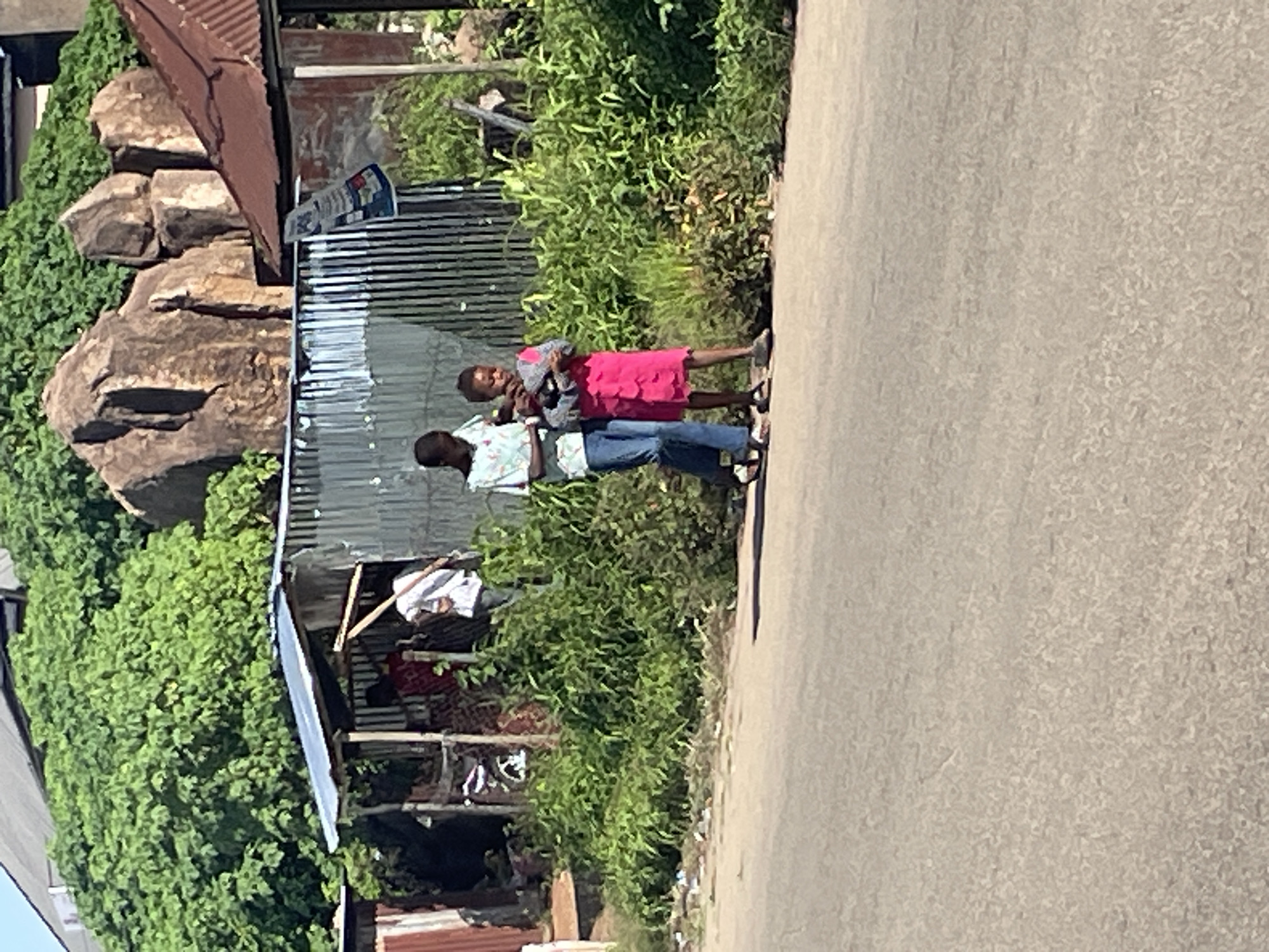 In Jos, Nigeria, two children walk along a paved road next to a corrugated metal structure with an open front, possibly a makeshift shop or shelter. Lush green vegetation grows behind the structure, with large rocks and trees visible further in the background. The scene suggests a semi-urban or rural setting.