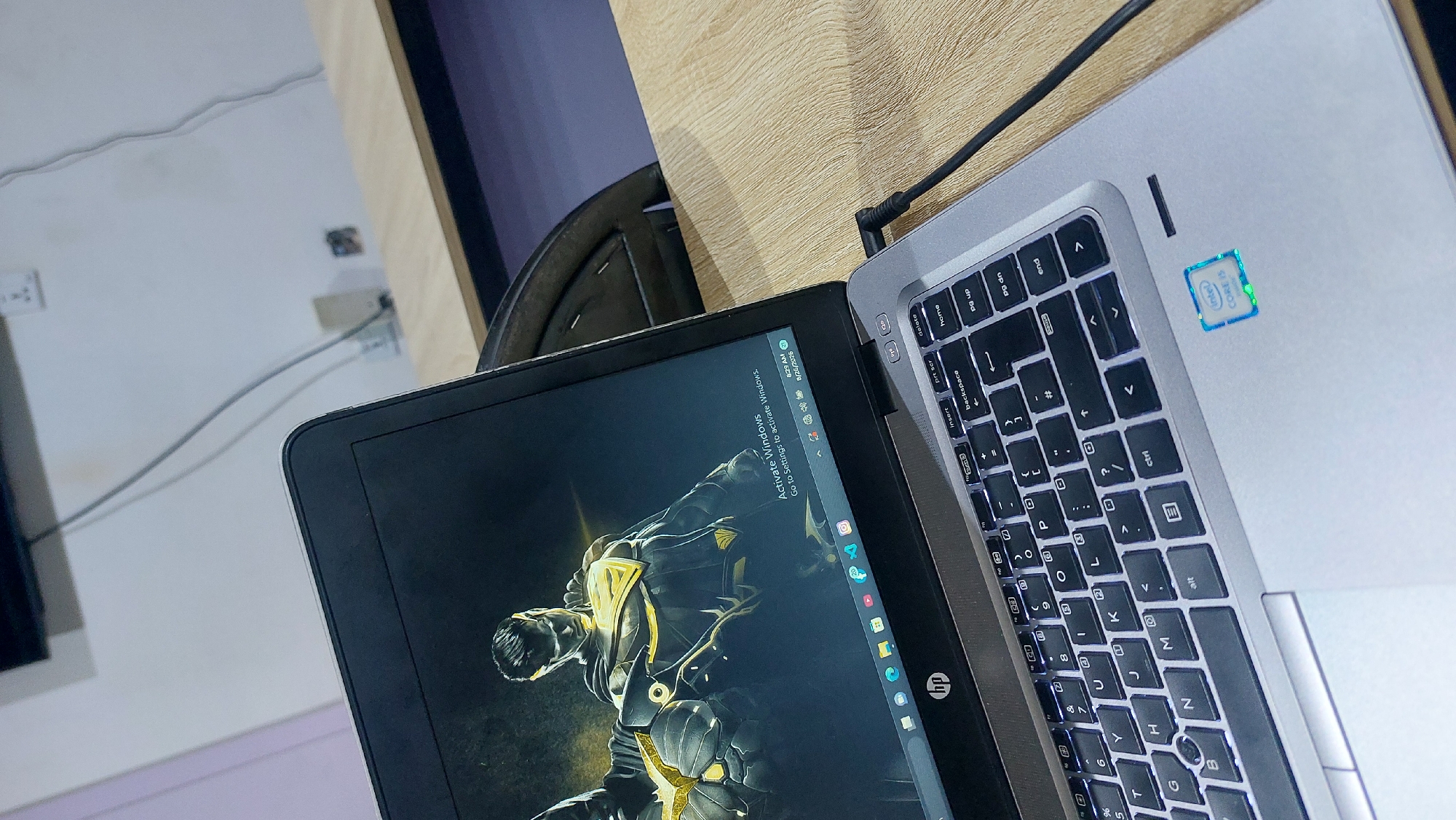 The image displays a close-up, eye-level view of a silver HP laptop open on a light brown wooden desk. The laptop's screen shows a dark wallpaper featuring two comic book characters, one in the foreground wearing a black and gold suit with a cape, and another partially visible character behind him. In the lower right corner of the screen, there's a watermark indicating "Activate Windows" and the system time is 8:29 AM on 8/26/2025. The keyboard is illuminated with white backlighting. A black power cable is plugged into the right side of the laptop. To the right of the keyboard, on the laptop's palm rest, is a blue and green "Intel Core i5" sticker. The background is blurred, showing a white wall with electrical outlets and a dark chair. The overall scene suggests an indoor office or study environment. There is no identifiable information to suggest this scene is located in Jos, Nigeria.