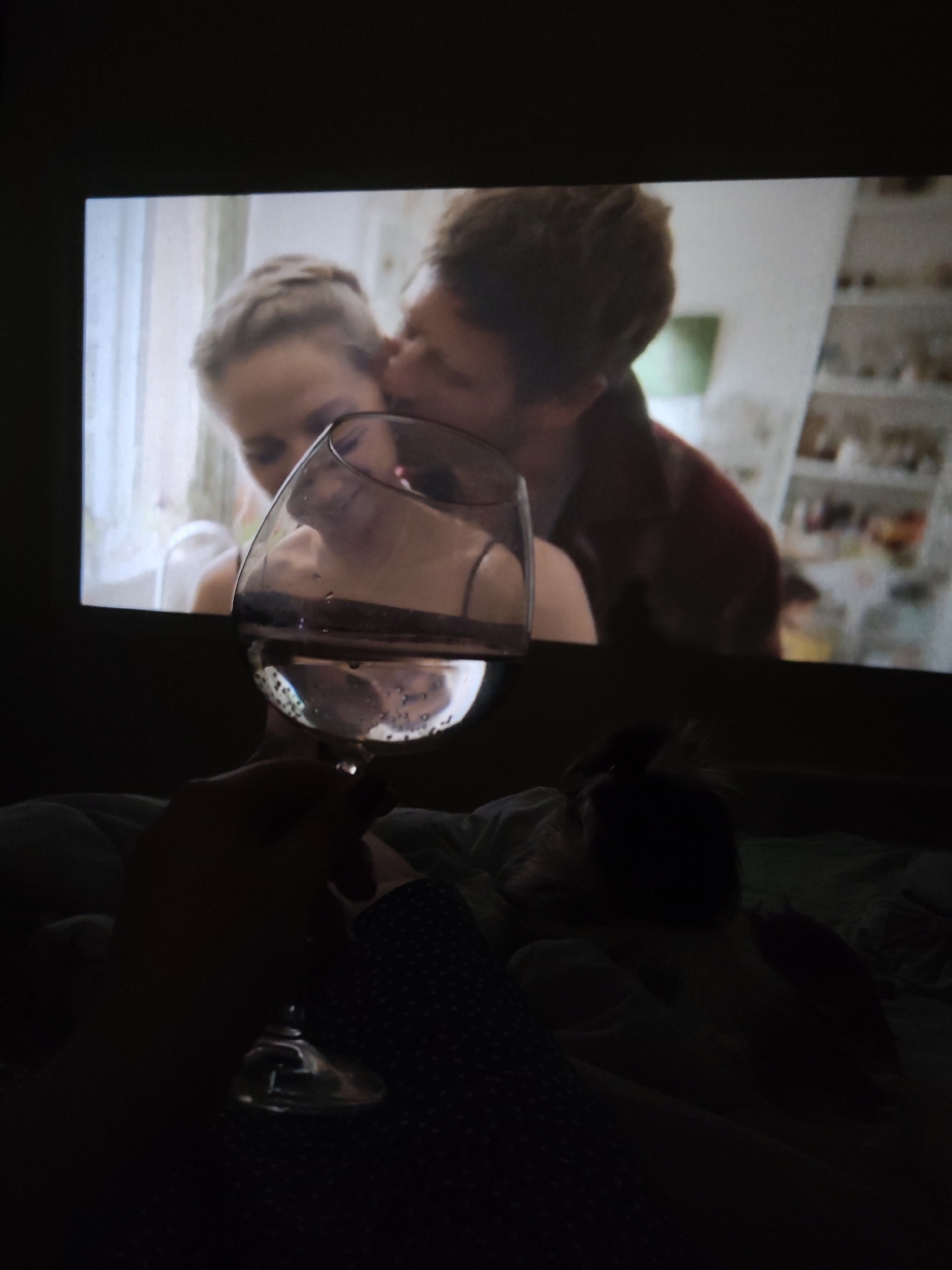 The image depicts a dark room with a projection screen displaying a scene of a man kissing a woman's cheek. A hand is holding a wine glass filled with liquid in the foreground, obscuring part of the screen. A dark shape, possibly a pet, is lying in the bed or on a surface below the glass. The location and city of Izhevsk, Russia cannot be confirmed based on the visual information provided in the image.