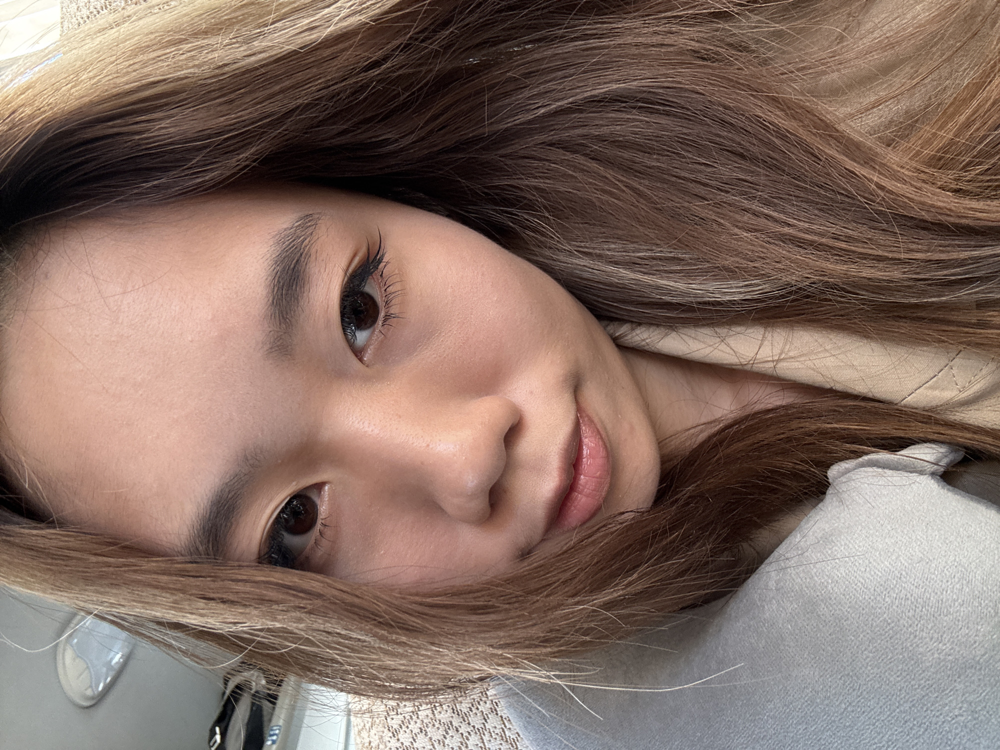A close-up, slightly angled portrait captures a young Asian woman with light brown hair and dark eyes. Her face is oriented to the viewer's left, with strands of hair framing her features. She has well-defined eyebrows, long eyelashes, and smooth skin. Her lips are a natural pink shade. The background is blurred, but elements of a light beige or gray fabric are visible, suggesting clothing or upholstery. A glimpse of what appears to be a light-colored wall is present on the far left. The image does not provide information about the city of Wuwei, China, or any specific location.