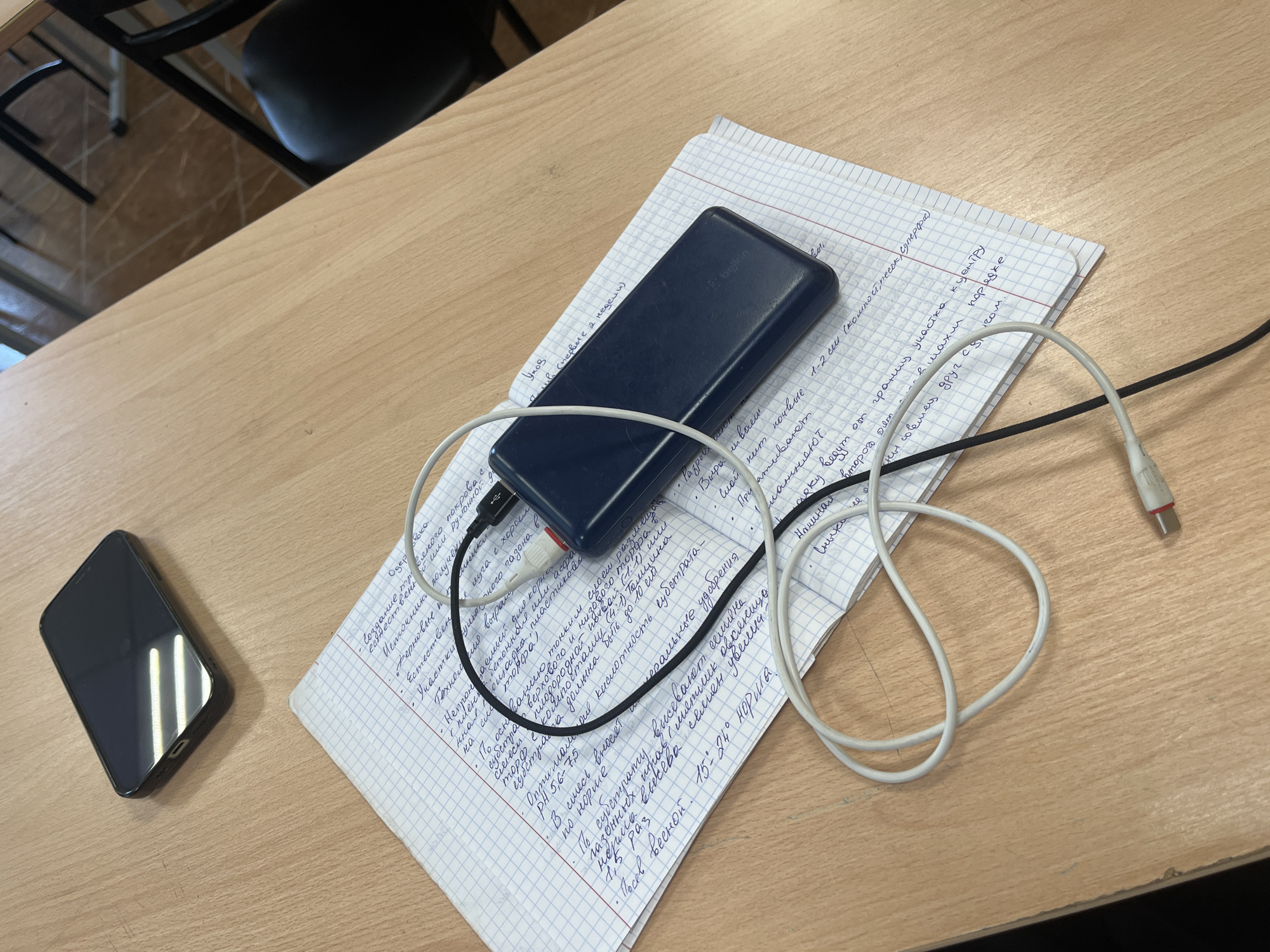 Here's a detailed description of the image:
**Scene:** A desk with a notebook containing handwritten notes in Russian, a smartphone, a portable charger (power bank), and charging cables.
**Objects:**
* **Notebook:** Open, with handwritten notes in Russian covering both pages. The paper is graph paper.
* **Portable charger:** Dark blue/black, resting on top of the notebook.
* **Cables:** Two cables are connected to the power bank. One is white with a USB-A connector at the end. The other is black with a USB-C connector at one end and a USB-A connector at the other.
* **Smartphone:** Black, lying face-up on the desk to the left of the notebook. The screen is dark.
**Environment:**
* The desk has a light-brown wood-grain surface.
* A chair is visible in the background (likely a school desk or office chair).
**Location:** There is nothing to indicate that this image was taken in Saint Petersburg, Russia.