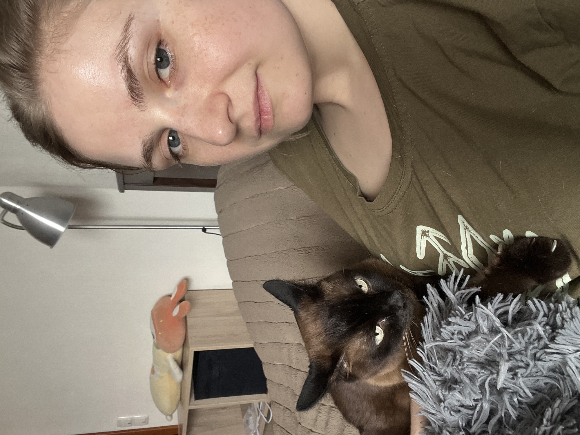 The image is a selfie of a fair-skinned woman with freckles and blue eyes. She wears a green t-shirt and has a neutral expression. A Siamese cat lies next to her on a brown textured surface, likely a bed. The cat's dark brown face and light brown body are visible. Part of a gray fuzzy blanket or rug is near the cat. In the background, there is a white wall with a silver desk lamp and a cartoon plushie on a light wood shelf. A power outlet is visible near the floor. The city and location are not discernible from the given visual information.
