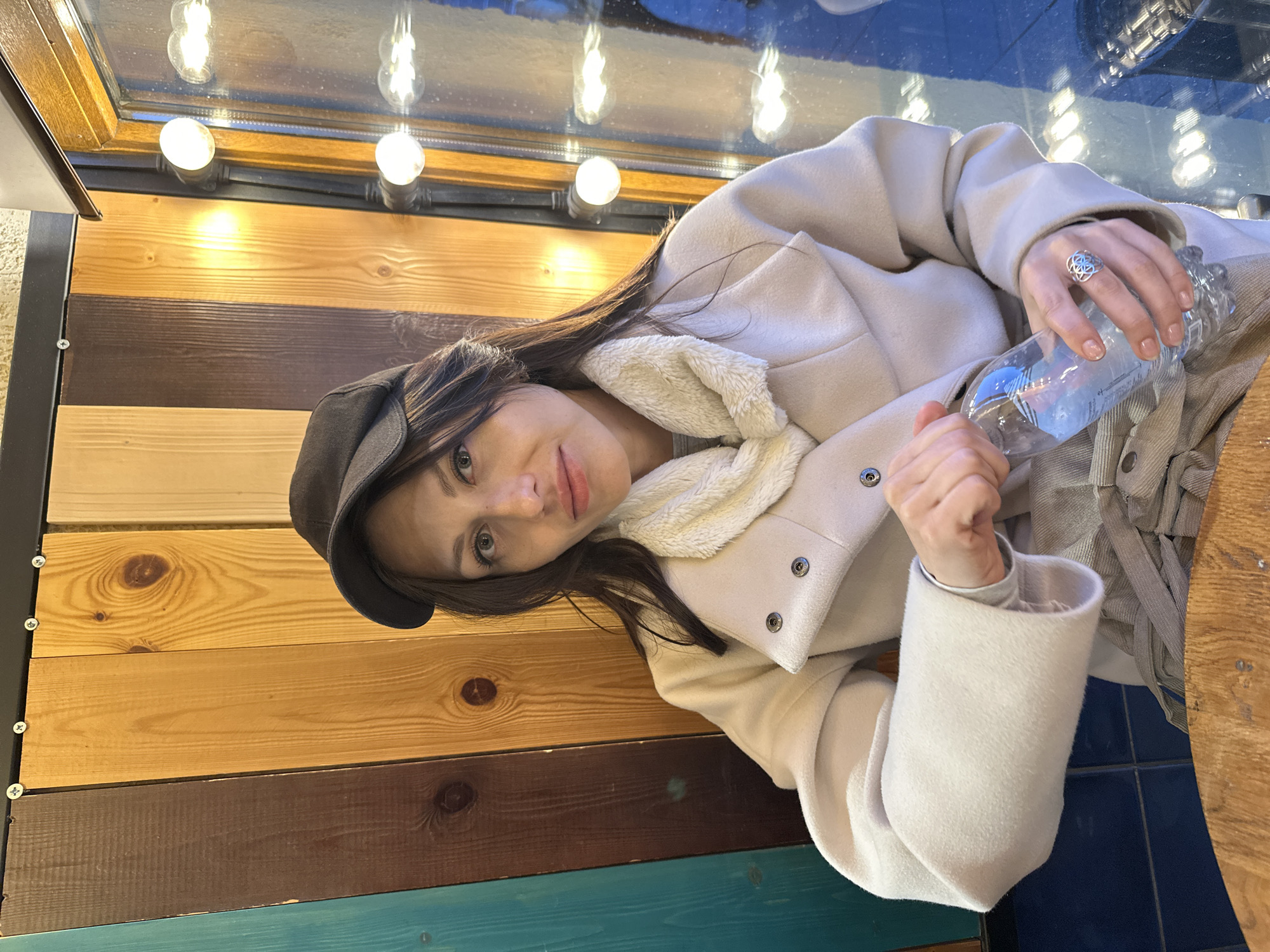 A woman wearing a beige coat with a fur-trimmed collar and a dark-colored cap is sitting indoors. She is holding a plastic bottle of water. She is wearing a ring on her right hand. The background is a wooden wall with different colored planks. Above her is a window with light bulbs. The floor is blue tile. The woman is sitting at a wooden table. The image might be in Saint Petersburg, Russia, but there are no specific details to confirm this location.