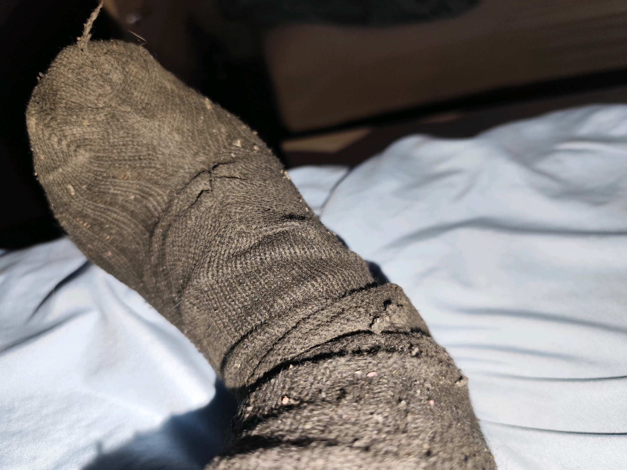 The image shows a close-up view of a person's foot wearing a dark gray, worn sock. The sock appears to be made of a knitted material and shows signs of significant wear, including fraying and loose threads, particularly around the ankle area. Small flecks of lint or debris are visible on the surface of the sock. The foot is resting on a light blue, wrinkled fabric, likely a bedsheet or blanket. The background is dimly lit and out of focus, with hints of brown furniture or a wall, and the overall lighting suggests it could be nighttime or an indoor setting with low light. No visible text is present in the image. The setting appears to be a domestic environment, possibly a bedroom. The worn state of the sock might suggest an informal or casual atmosphere. No specific activity or event is clearly depicted beyond the static presentation of the foot and sock.
